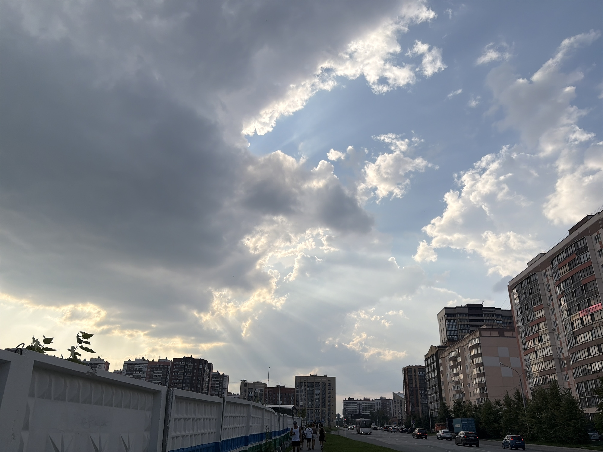 This is a landscape photograph of a street scene in Novosibirsk, Russia, captured during daylight hours under a cloudy sky with dramatic sunbeams. The image is dominated by a large, dark cloud bank on the left, partially obscuring the sky, with rays of sunlight breaking through from the right side of the frame. A row of multi-story apartment buildings lines the right side of the street, featuring a variety of architectural styles and colors, with many windows visible. Some of the buildings have balconies. A white concrete wall with a blue and green trim runs along the left side of the foreground, separating the sidewalk from what appears to be a utility area or construction site, with some sparse vegetation growing near the top of the wall. A paved road with traffic runs parallel to the buildings, with several cars and a bus visible. A small group of people, appearing to be pedestrians, are walking on the sidewalk in the middle ground, facing away from the camera. The overall atmosphere is one of an urban environment experiencing dynamic weather conditions.