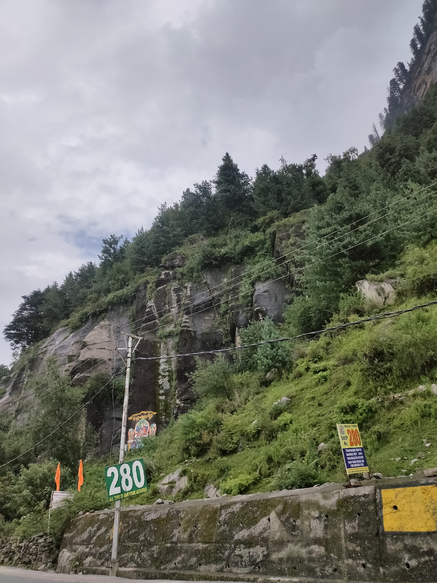 The image depicts a roadside scene in the mountainous region of Kulang, India. A steep, rocky cliff face covered in lush green vegetation rises dramatically on the right side of the frame. A cluster of tall, dark green pine trees grows along the ridge of the cliff. Power lines crisscross the scene, supported by wooden poles.
In the foreground, a concrete retaining wall borders a paved road. On the left side of the road, a wooden pole displays a green sign with white numbers "280" and text below that reads "ALL TYPE OF ADVENTURE ACTIVITIES". To the left of this pole, two bright orange flags on poles are visible. Further up the cliff face, near the pole, a colorful religious mural is painted, depicting deities.
On the right side of the frame, closer to the road, another pole supports a yellow and blue sign with "HIMACHAL TOURISM" at the top, followed by "SHOP NO. 280" and details about tourism rates. The sky is overcast with a grayish, cloudy appearance. The overall impression is a scenic, yet functional, roadside view in a mountainous area.