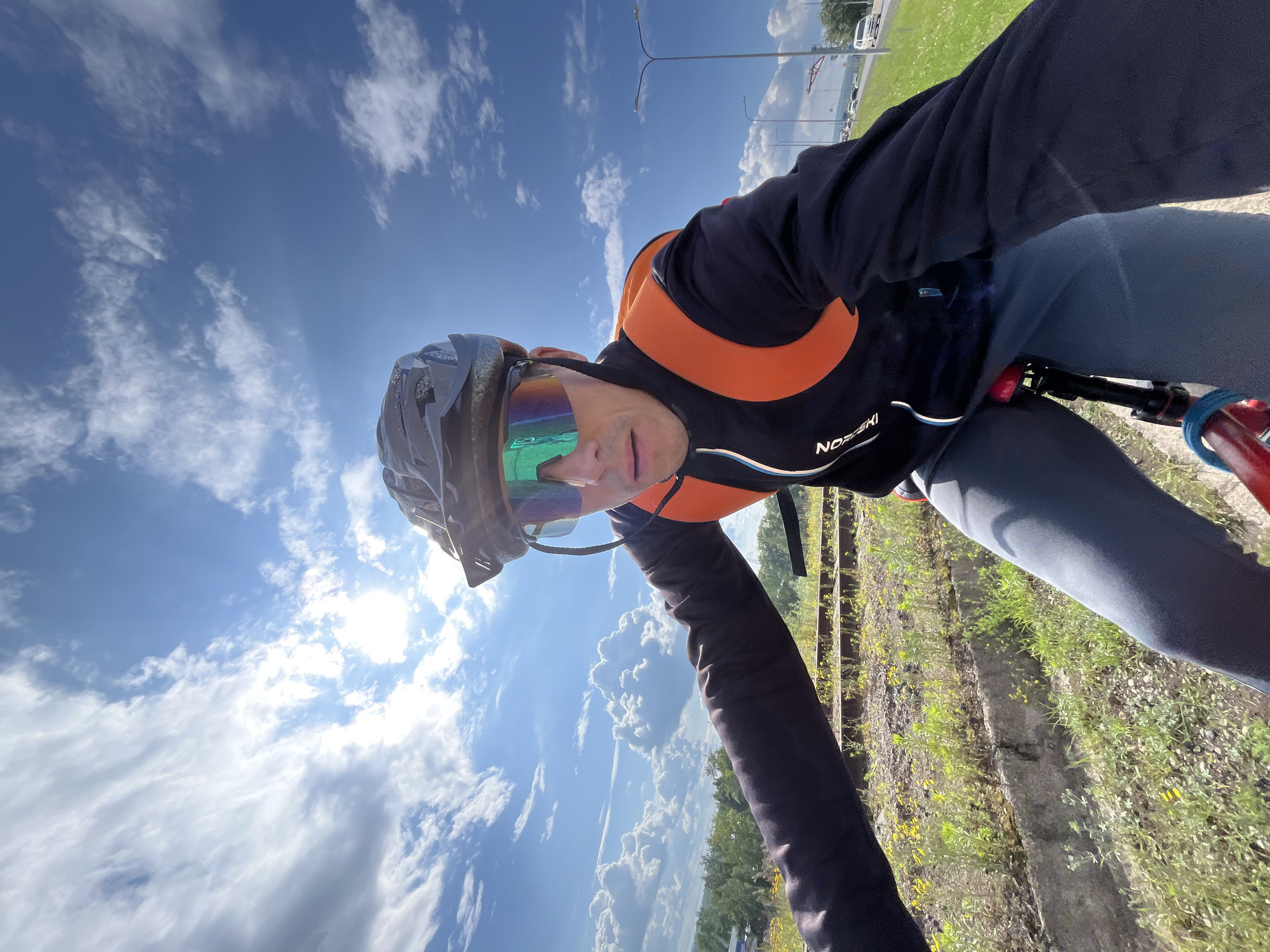 A man wearing a black helmet and neon green cycling glasses is riding a bicycle. He is dressed in a black long-sleeved jacket with an orange vest over it and grey pants. The man is looking directly at the camera. The background shows a bright blue sky with scattered white clouds, green vegetation, and a road with some traffic in the distance. This scene is likely in or around the city of Chaikovsky, Russia, given the presence of outdoor recreational infrastructure and potential proximity to common routes.