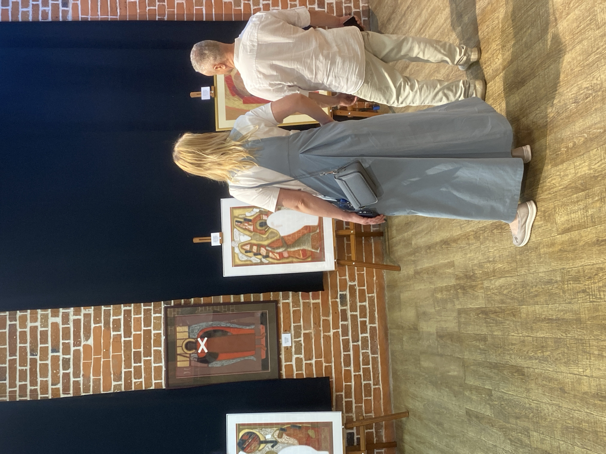 An indoor scene at an art gallery in Yekaterinburg, Russia. A middle-aged, grey-haired man wearing a white shirt and tan pants is viewing artwork. A woman with blonde hair wearing a light blue dress and a white shirt is also viewing artwork. The walls are a combination of brick and black fabric. Three framed paintings are visible on easels. The floor is wooden.