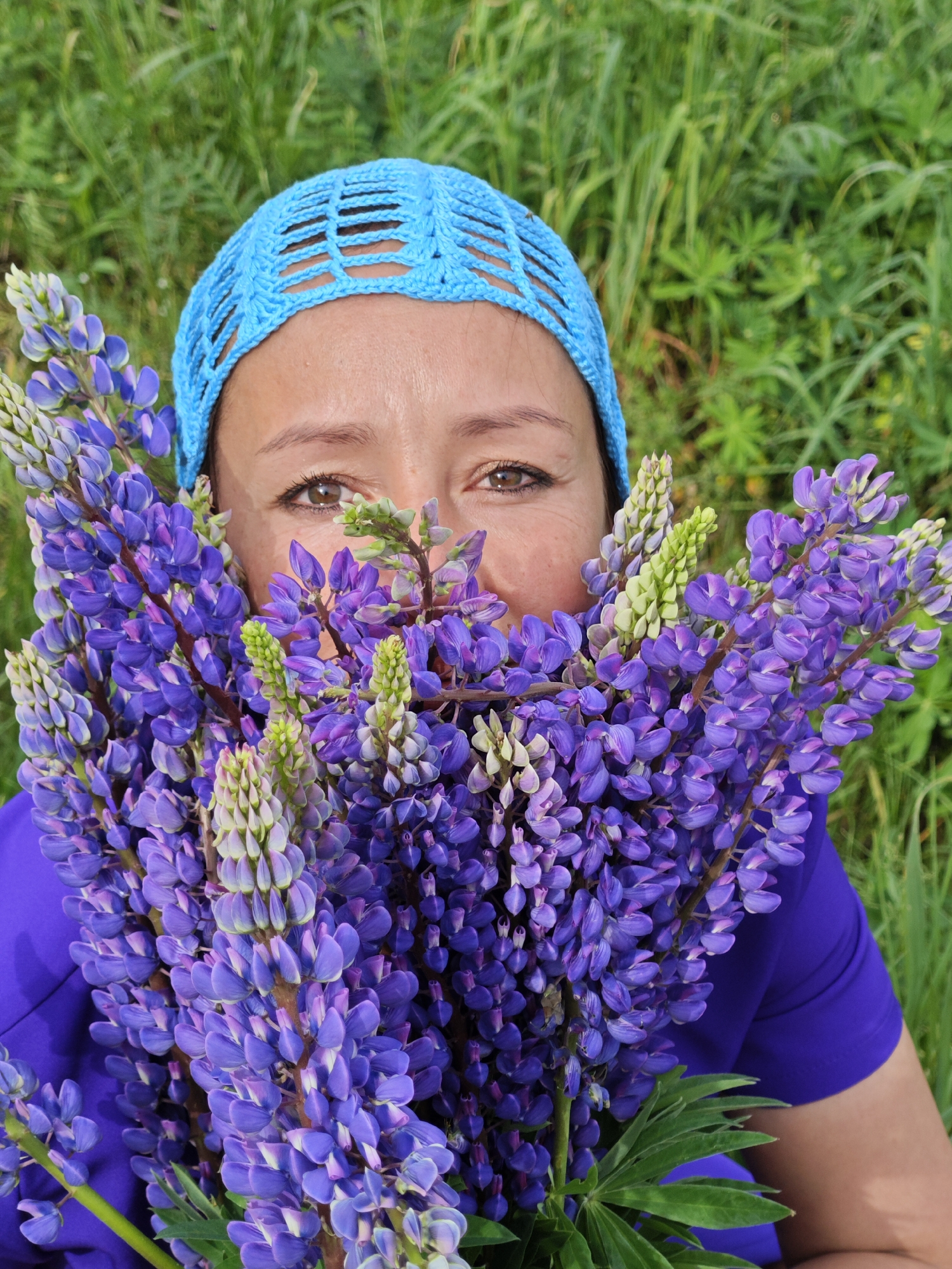 A woman with a blue crocheted cap is holding a large bouquet of purple lupine flowers in front of her face in an outdoor setting. Only her eyes and forehead are visible above the flowers. She is wearing a blue shirt. The background is blurry green grass. The location may be near the city of Chaikovsky, Russia, as the city is known for its lupine flowers.