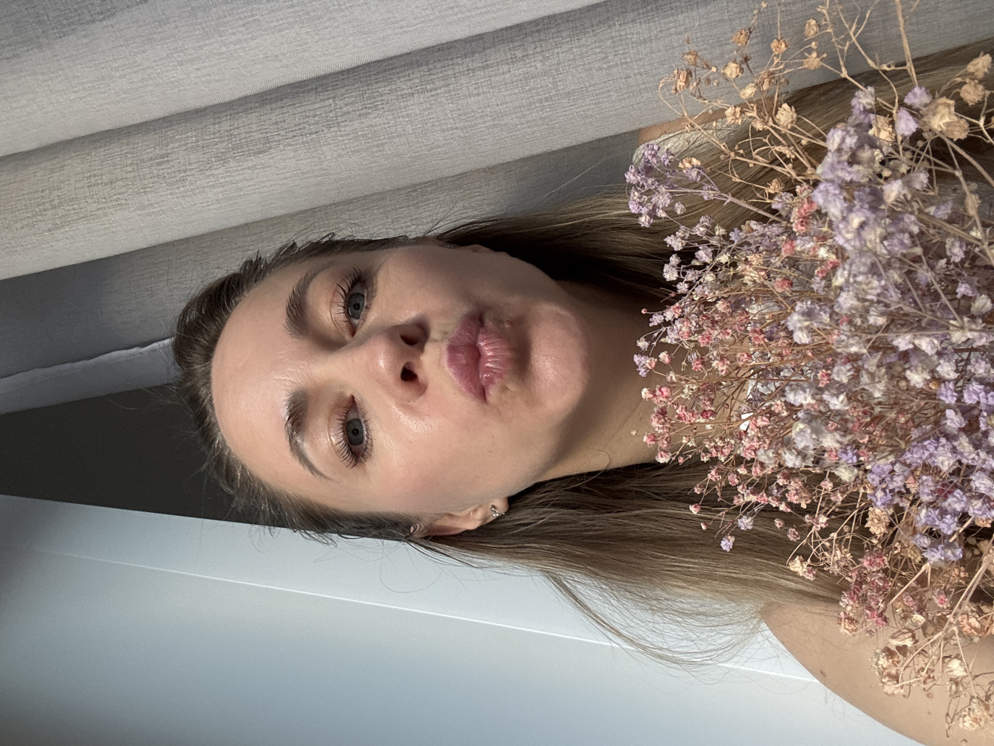 A close-up, portrait-style photograph depicts a woman posing with a bouquet of dried flowers. The image is oriented vertically, showing the woman's face from the nose up to her forehead, and her shoulders, while her long, light brown hair cascades around her. Her head is tilted slightly.
The woman has fair skin, light-colored eyes (appearing blue or grey), and prominent eyelashes. Her eyebrows are well-defined. She is making a "kissing face" with her lips pursed, which are full and have a pinkish hue. A small, silver-colored earring is visible in her left ear.
To the right of the woman, a dense cluster of dried, delicate flowers is prominently featured. These flowers are small and have faded hues of light purple, dusty pink, and beige/brown, indicating they are dried baby's breath or a similar variety. The flowers are arranged closely to her face and hair.
The background consists of two main elements. In the upper portion, behind the woman's head and the flowers, there is a light grey or beige textured fabric, possibly part of blinds, curtains, or a headboard, showing horizontal lines. The lower portion of the background, beneath her chin and hair, is a smooth, plain light grey or white surface.
The lighting is soft and even, suggesting an indoor setting. There is no visible text in the image. The overall impression is a posed, artistic shot focusing on the woman's expression and the rustic charm of the dried flowers in Chaikovsky, Russia.