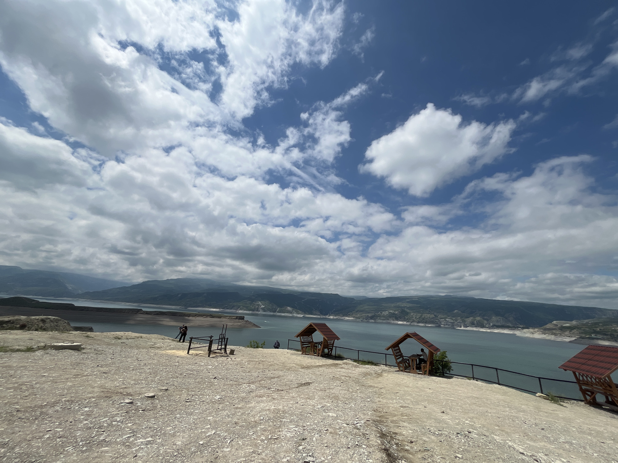 The image shows a wide view of a reservoir or lake in Chirkei, Russia, under a sky with scattered white clouds. In the foreground is a dirt and gravel area with a slight slope. Three wooden gazebos with red-tiled roofs are positioned along the edge of the area, overlooking the water. One person is visible sitting inside one of the gazebos. Farther to the left, two people stand next to what appear to be exercise equipment. In the background, hills and mountains are seen across the body of water.