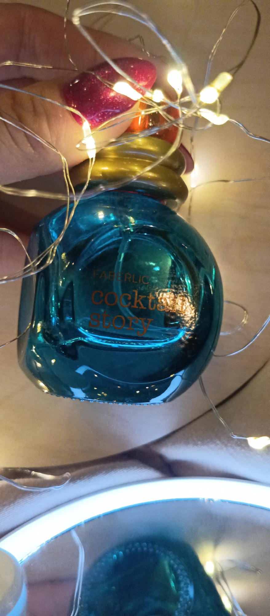 A close-up shot reveals a blue glass bottle labeled "Faberlic Cocktail Story" held by a person with a burgundy glittery nail polish. The bottle is surrounded by thin, silver wire lights that emit a warm glow. The background appears to be a soft, light-colored fabric or surface. No location information is available.