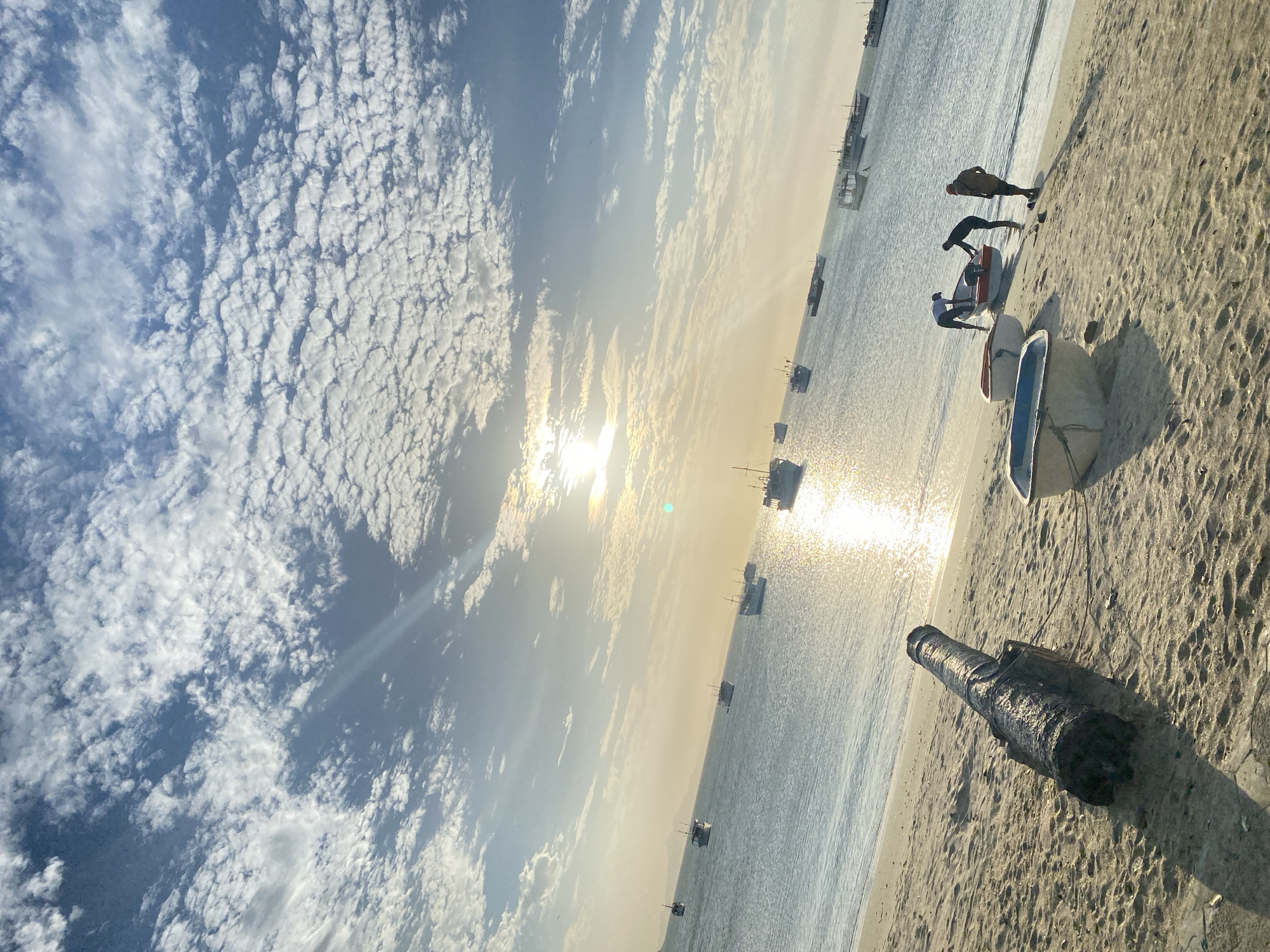 The image depicts a beach in Juan Griego, Venezuela. The sky is filled with small, puffy clouds. The sun is shining, creating a glare on the water. Several boats are anchored in the sea. On the beach, there are several small fishing boats, and three people are near the boats, appearing to be working on them. A cannon is present on the sand in the foreground.