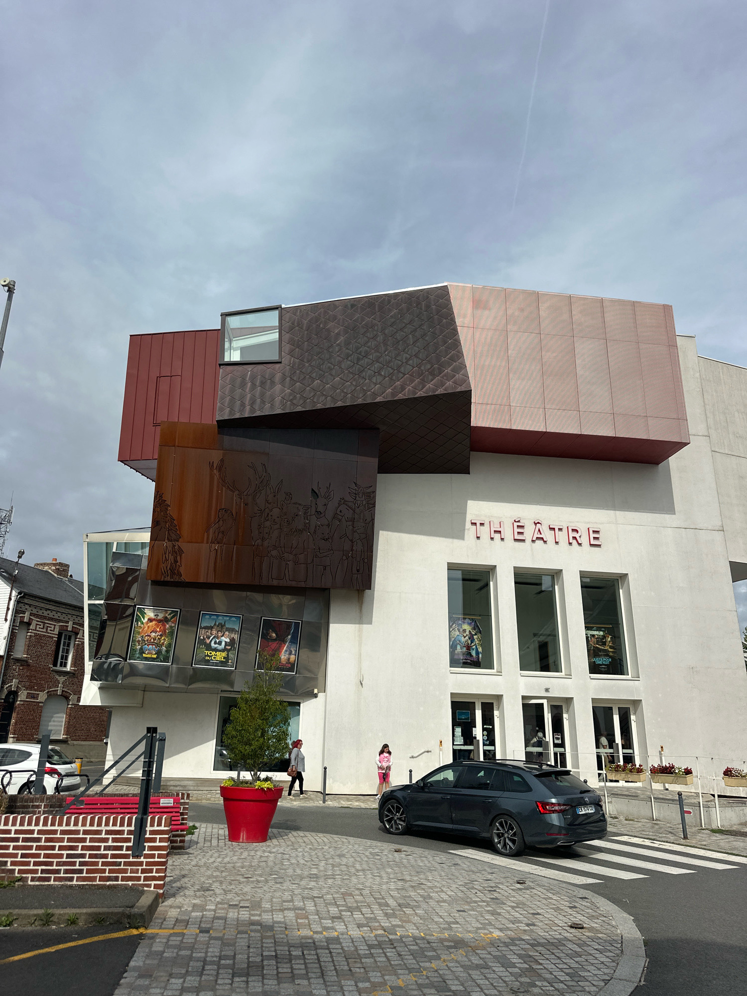 The image depicts the exterior of a theater in Roye, France, on a cloudy day. The building features a modern architectural design with angular, overlapping sections clad in various materials, including copper-toned metal and perforated red panels. The name "THÉÂTRE" is prominently displayed in large, light pink letters above three large windows on the main white facade. Below these windows are entrances.
In front of the theater, a dark grey station wagon is driving across a pedestrian crossing. To the left, a white car is parked near a brick structure with a red bench. A large red planter with a small tree is situated on the cobblestone sidewalk. Three movie posters are displayed on the building's facade, to the left of the "THÉÂTRE" sign. A person in a pink jacket and white pants is standing near the entrance, and another person in a grey jacket and black pants is walking away from the building. The sky is overcast with a faint contrail visible. The street is paved with cobblestones and asphalt, with yellow painted lines indicating traffic lanes.