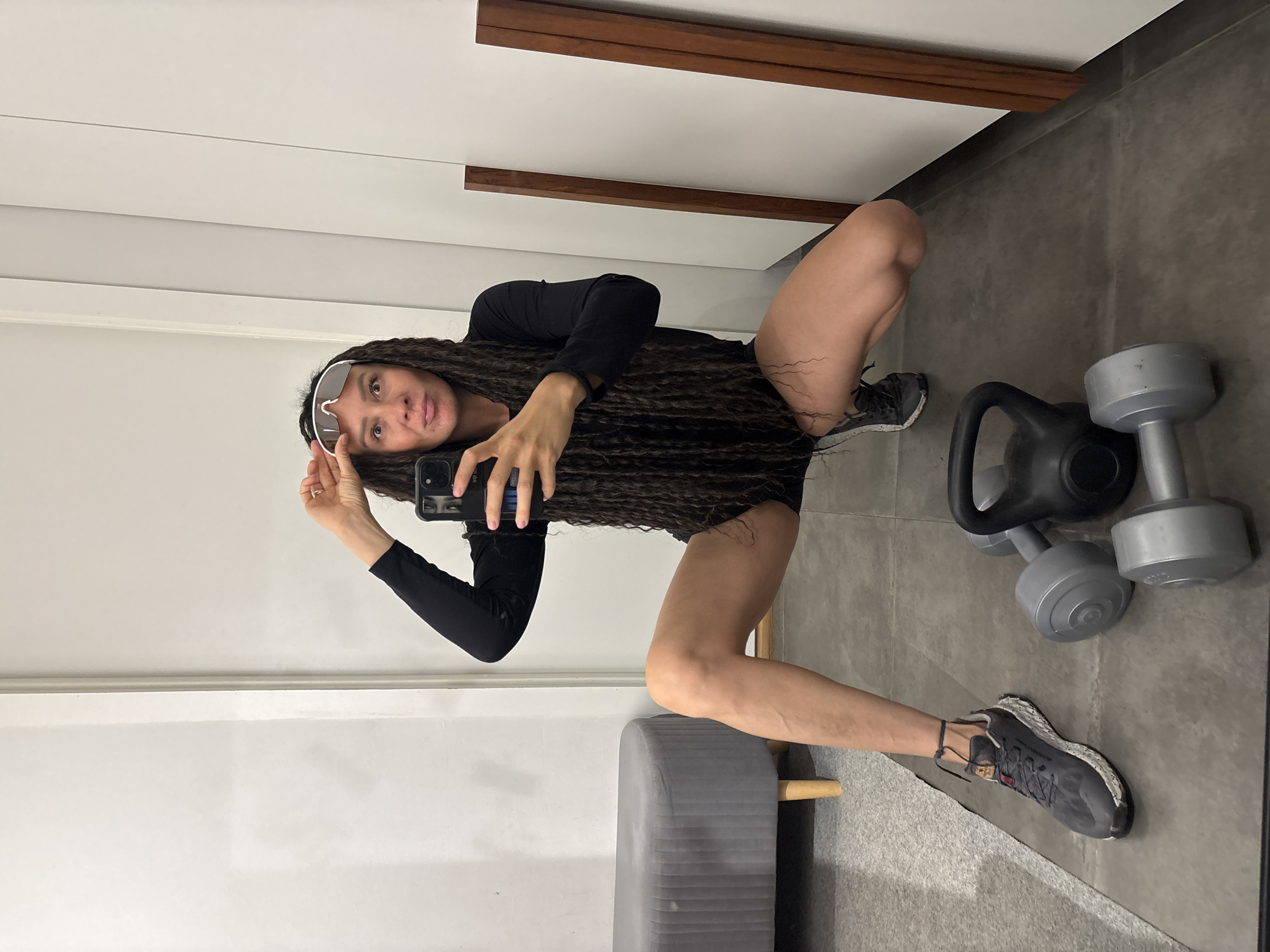 A woman with long, dark, braided hair is captured in a mirror selfie within an indoor setting, likely a home gym or fitness area. She is in a low crouch or squat position, looking directly at the camera. She wears a black long-sleeved top, black shorts, and dark grey athletic shoes with speckled white soles. White-rimmed athletic sunglasses are pushed up onto her forehead. She holds a black smartphone in her right hand to take the picture.
The background consists of a grey tiled floor and a white wall or cabinet with two horizontal dark brown wooden accents. To the woman's right, a black kettlebell and several grey dumbbells of varying sizes are visible on the floor. To her left, part of a grey upholstered bench or stool with light wooden legs is visible. The lighting is even, suggesting an indoor environment. Her expression is neutral, with her tongue slightly visible between her lips. There is no discernible text. The image does not provide specific cues to the suggested location of Chaikovsky, Russia.