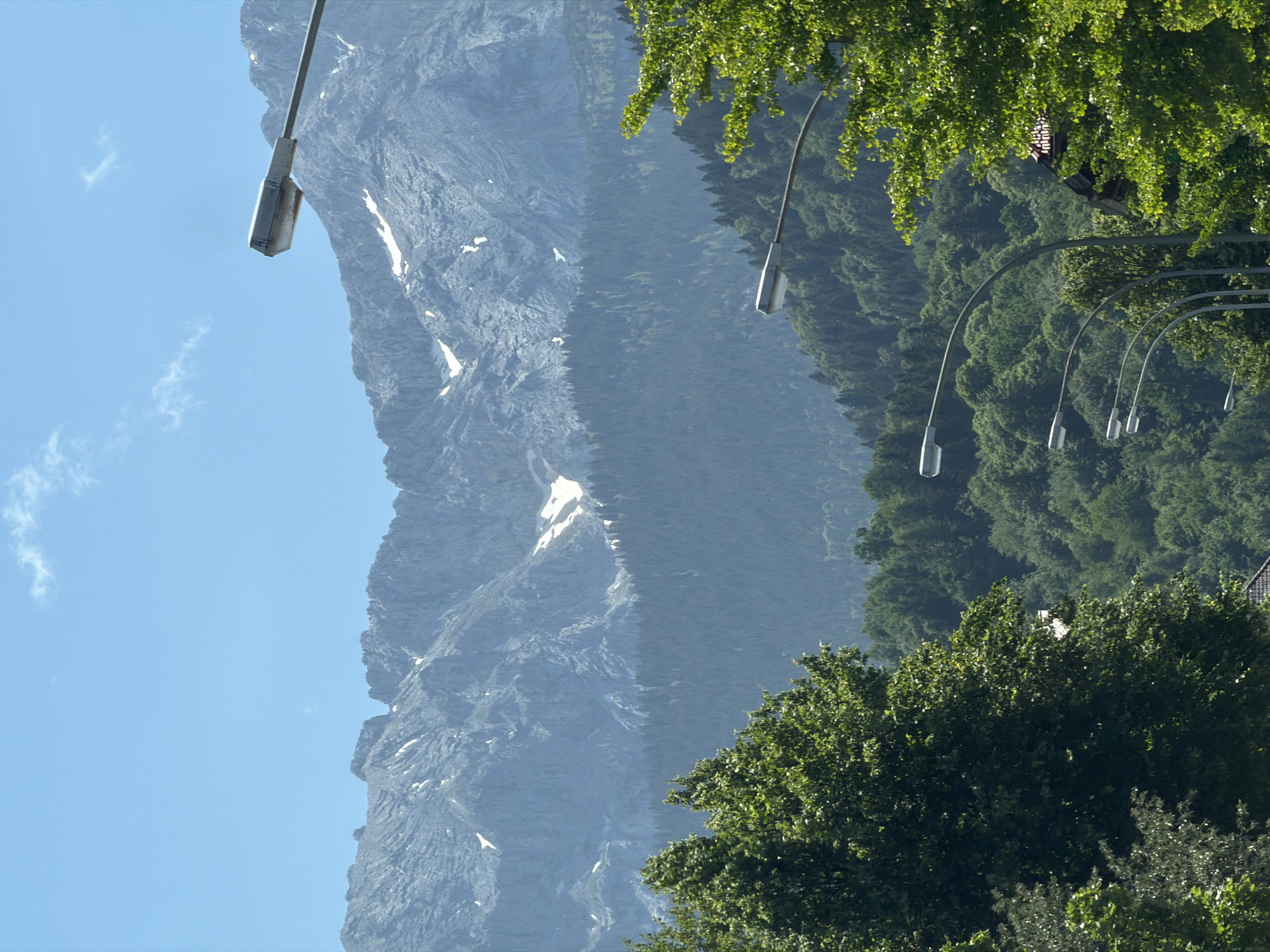 The image captures a scenic view of the Zugspitze mountain range, with lush green trees in the foreground and a clear blue sky with a few wispy clouds above. Several streetlights are visible, arching over the scene, suggesting a road or pathway. The mountains display rugged, rocky terrain with patches of snow, indicating a high altitude. The dense forest covering the lower slopes of the mountains adds to the natural beauty of the landscape. A small building or roof structure is partially visible through the trees on the right side of the image. The overall impression is one of a serene and majestic natural environment on a bright, sunny day. The presence of streetlights and the viewpoint suggest this scene is likely from an elevated position overlooking the town of Garmisch-Partenkirchen, Germany, offering a panoramic vista of its surrounding natural landscape.