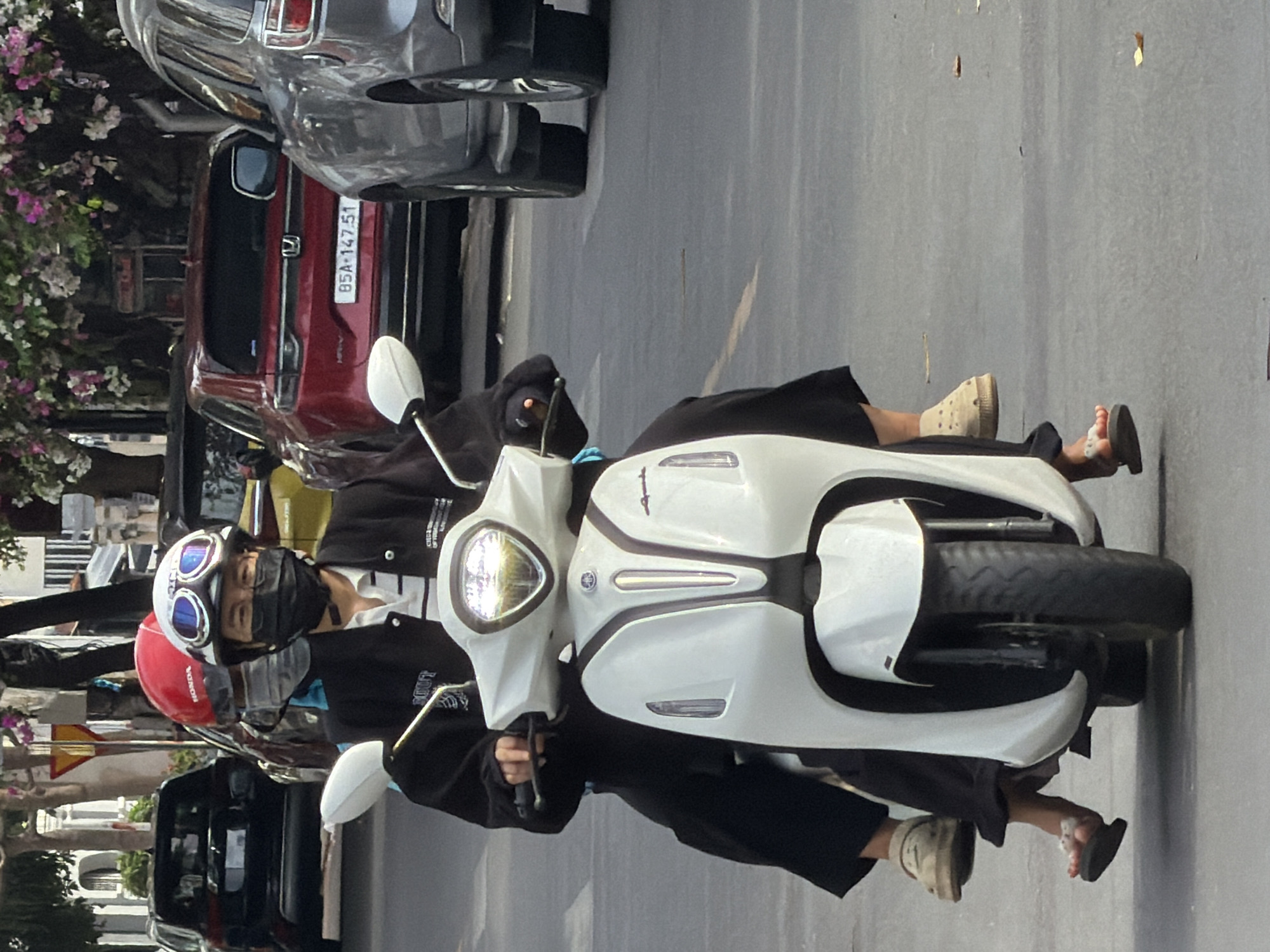 Two individuals are riding on a white scooter on a city street. The person in the front, driving the scooter, is wearing a black jacket over a striped shirt, a black face mask, and a white helmet with goggles. The person behind is wearing black pants and a light-colored shoe and is partially obscured by the driver. The scooter has a prominent white body with black accents and a clear windshield. The street appears to be paved with asphalt. In the background, there are several vehicles, including a red Honda SUV with a license plate that reads "85A-147.51," a gray SUV, and a black SUV. Trees with pink and white flowers are visible along the side of the road, suggesting a tropical or subtropical urban environment. The lighting indicates it is daytime, likely with sunlight. The presence of the license plate format and the vehicles suggest the location is in Vietnam, possibly Nha Trang, given the context.