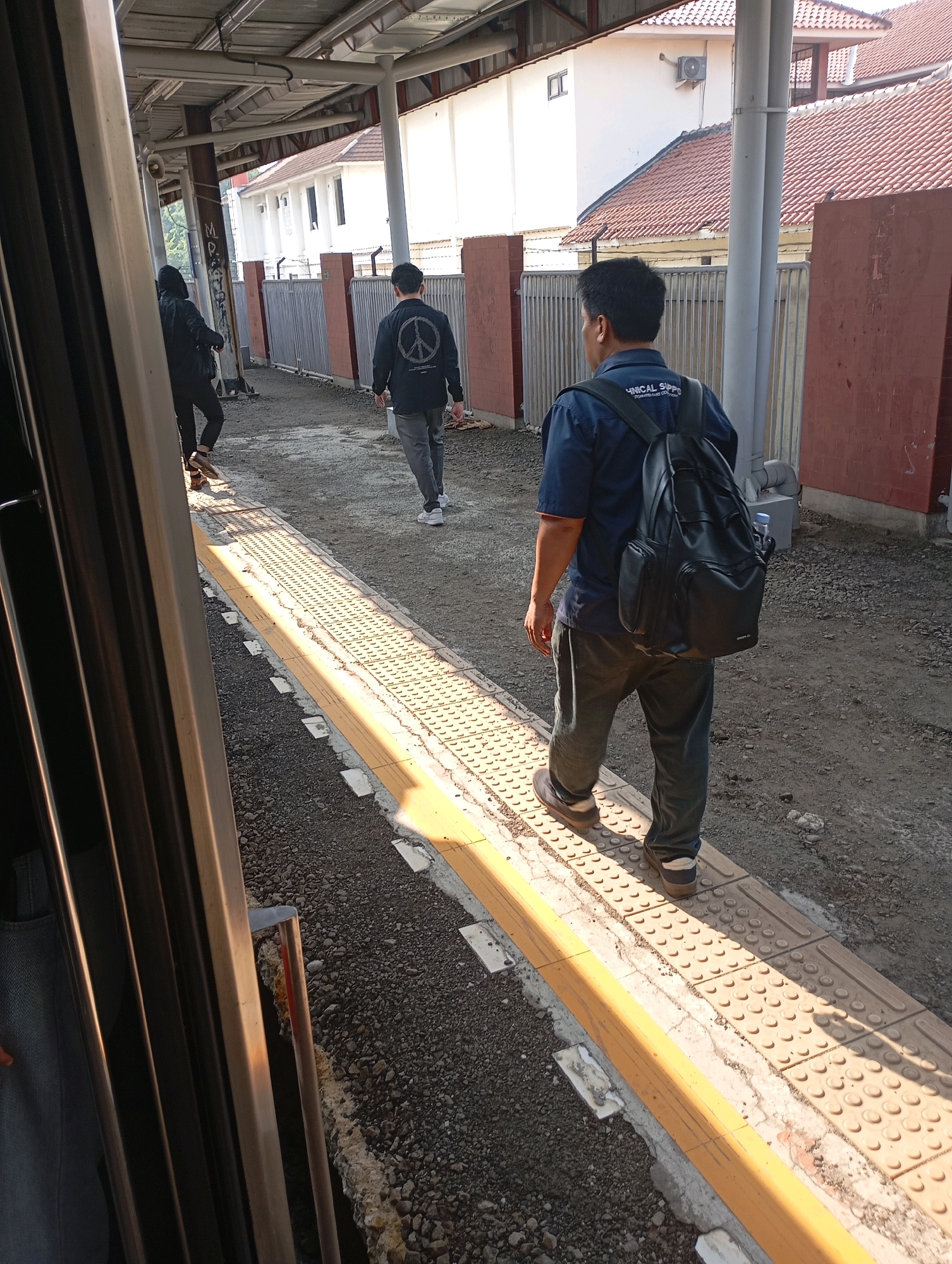 The image shows a train station platform in West Jakarta, Indonesia, on a sunny day. In the foreground, a train car's exterior is visible on the left. On the platform, a man wearing a blue short-sleeved shirt and dark pants, carrying a black backpack, walks away from the camera. Another person in a black hooded jacket and dark pants is walking in the same direction further down the platform. The platform has tactile paving and a yellow safety line. The background shows a building with a tiled roof and a metal fence. The ground is covered in gravel and dirt.