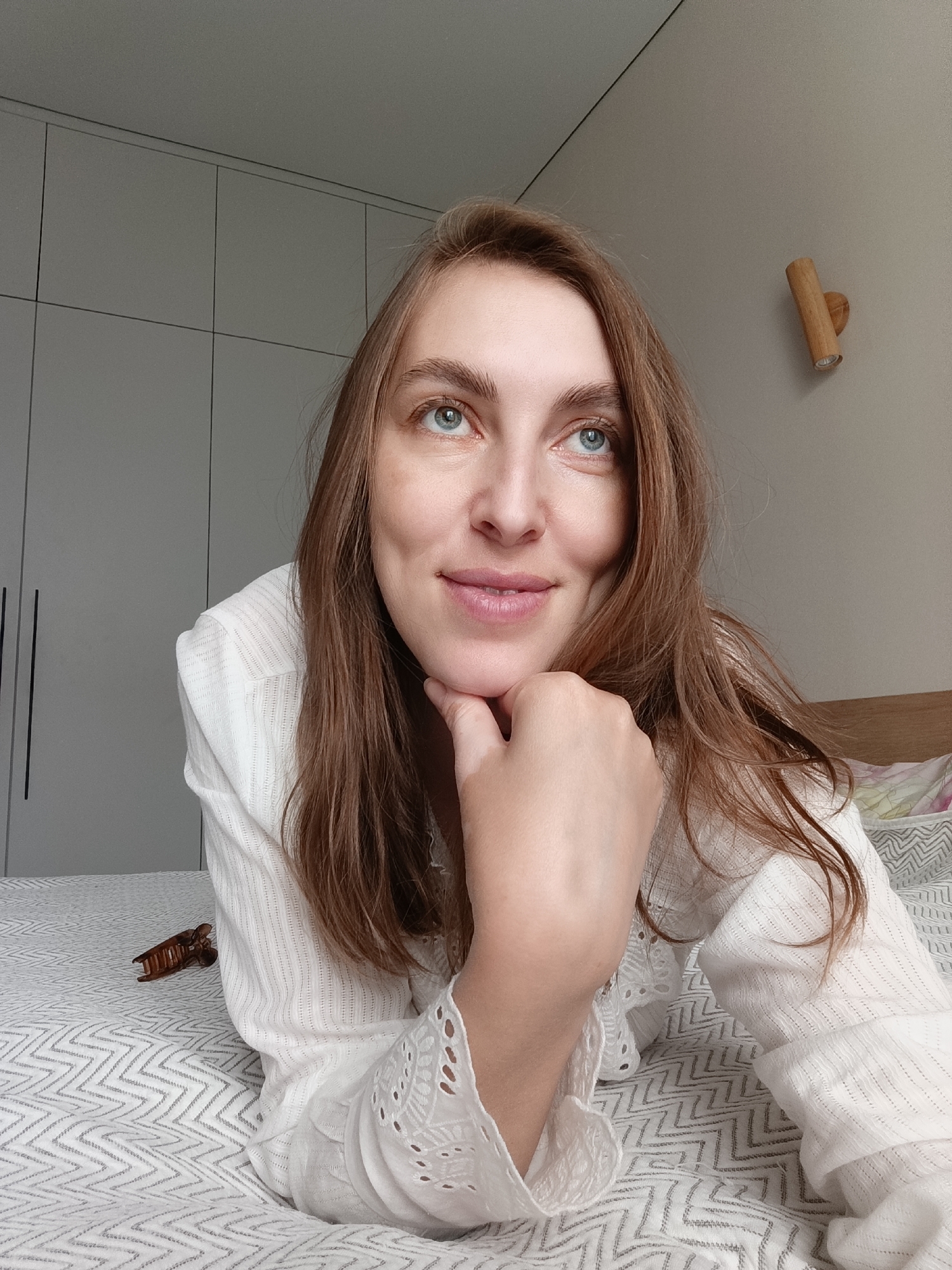 A woman with shoulder-length brown hair and blue eyes rests her chin on her hand while lying on a bed with a patterned gray and white cover. She wears a white, long-sleeved shirt with lace detailing. A brown hair clip is visible on the bed next to her. Behind her, a gray wardrobe cabinet stands against a light gray wall, where a wooden wall lamp is mounted. The image was taken in the city of Chaikovsky, Russia.