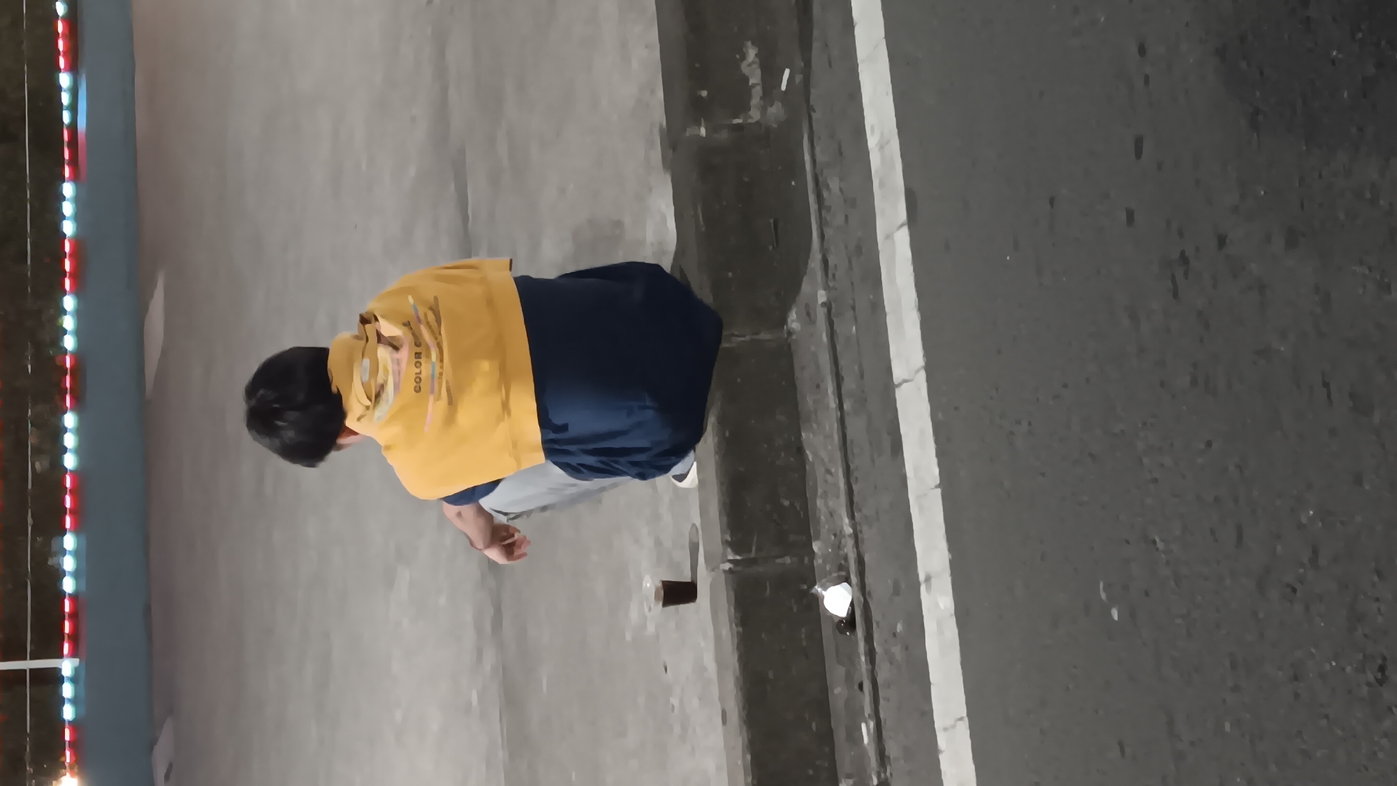 A person is seen from behind, squatting on a concrete curb at night in what appears to be an urban setting. The individual is wearing a yellow hoodie draped over their shoulders and a dark blue t-shirt underneath, with light-colored pants and white sneakers. A small, clear plastic cup containing a dark liquid sits on the curb next to them. In the foreground, the asphalt road is visible, marked with a white line. In the background, a row of colorful lights is illuminated against a dark wall or structure. The scene is dimly lit, suggesting it is nighttime. The environment appears to be an outdoor public space in South Jakarta, Indonesia, possibly a sidewalk adjacent to a road.