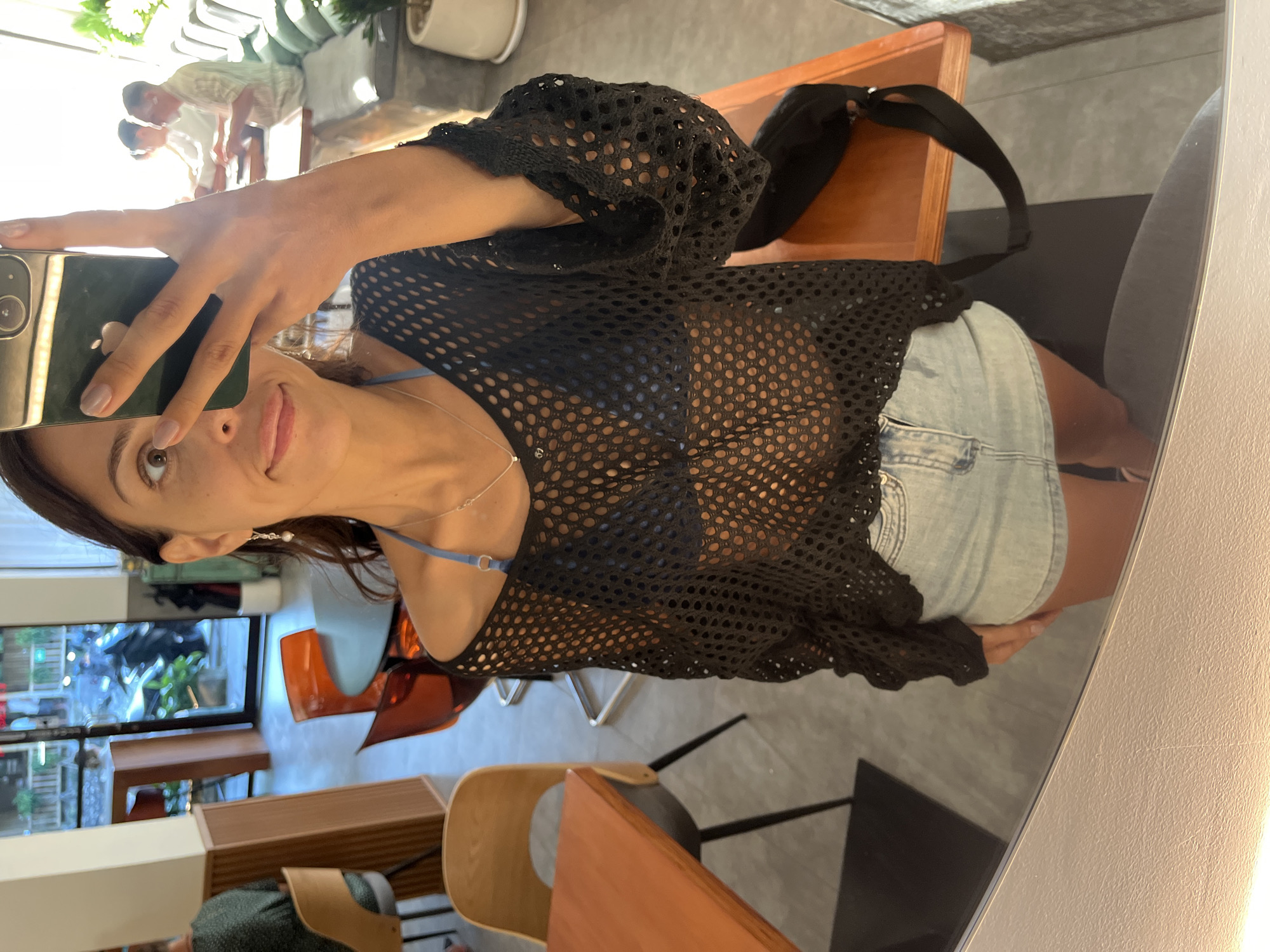 This is a mirror selfie taken by a woman in what appears to be a modern indoor setting, likely a café or restaurant. The woman, with dark hair and light-colored eyes, is looking towards the camera with a subtle smile, holding a smartphone with a dark case that features a visible Apple logo.
She is wearing a black long-sleeved mesh top over a light blue bikini top, paired with light-wash denim shorts. A delicate silver necklace and an earring with a pearl dangle are visible. Her nails are painted a light, neutral color.
The background, seen through the mirror's reflection, shows a bright, contemporary space. Two men in light-colored shirts are visible in the distance, appearing to be standing or interacting. Modern furniture, including wooden chairs, an orange plastic chair, and wooden tables, fills the space. A large window reflects lush green foliage outside, suggesting a warm and possibly tropical environment, consistent with Kuta Selatan, Indonesia. The floor is a light gray. The image captures a casual, daytime moment.