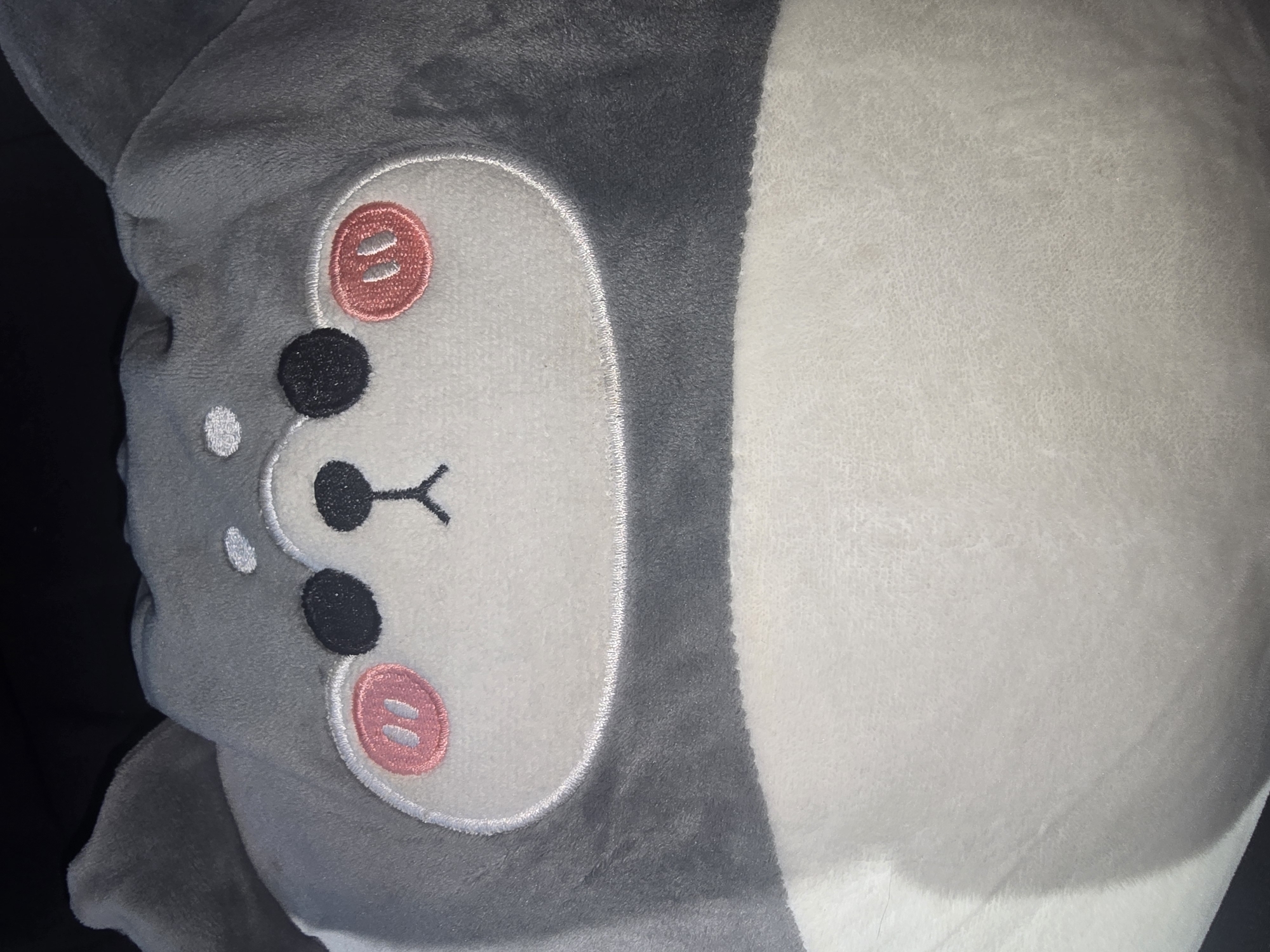 The image shows a close-up view of a plush toy, likely a dog or a similar animal, with a grey and white fuzzy exterior. The toy has embroidered features including two black eyes, pink circular cheeks with two vertical stitch marks each, and a small black embroidered nose and mouth. Its ears are pointed and grey. The background is dark and out of focus. There is no indication of the city of Turka, Russia, or any other specific location in the image.