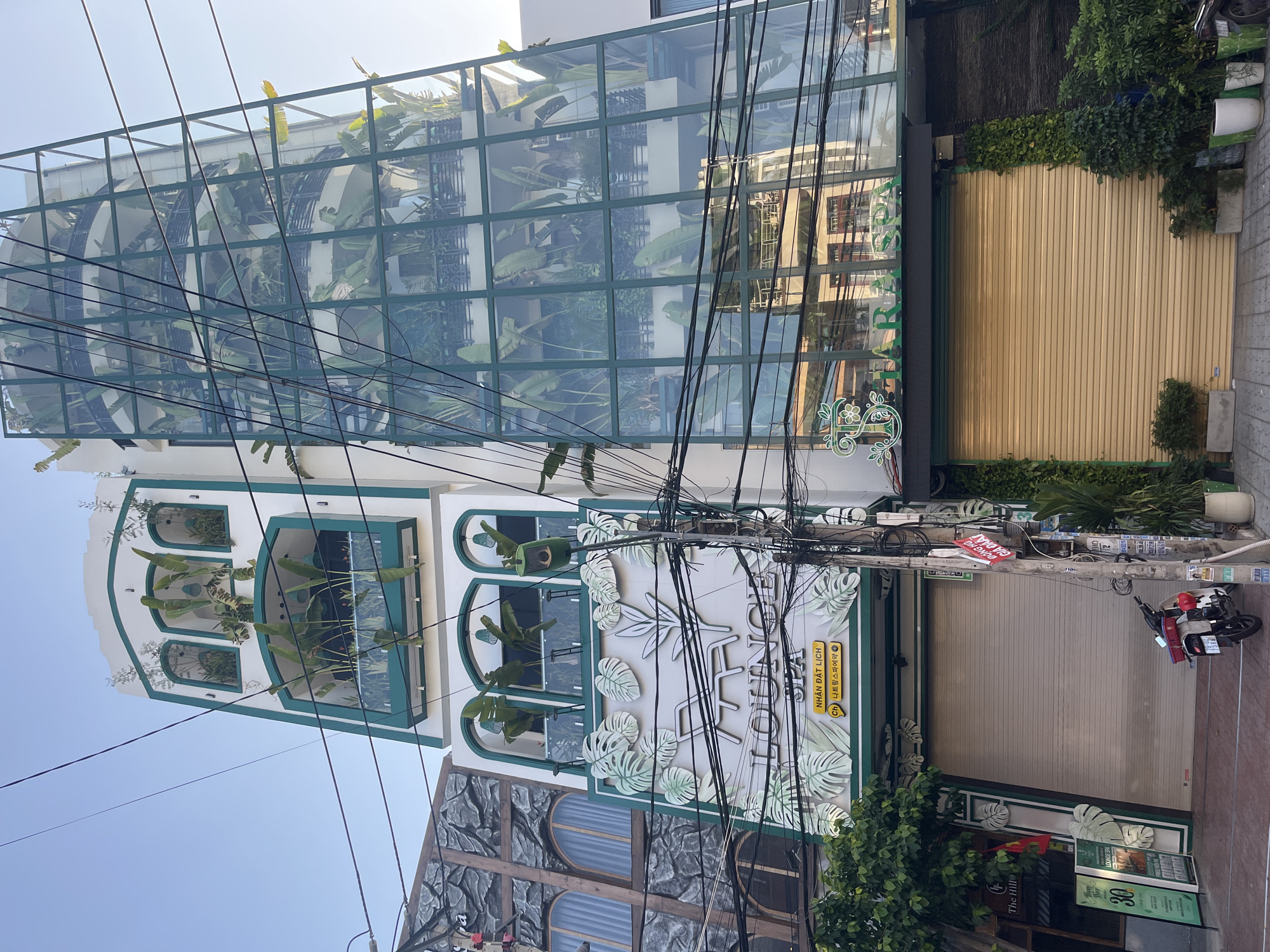 The image shows the exterior of two adjacent buildings on a street in Nha Trang, Vietnam. The building on the left is a white, multi-story structure with green trim. It features several arched windows on its upper floors, each containing potted plants. The lower facade of this building displays a large, white decorative motif of leaves and a chaise lounge, with the word "LOUNGE" prominently displayed in silver lettering. Below "LOUNGE", there are Vietnamese and Korean inscriptions that translate to "Nhân Dật Lịch" and "나트랑 스파예약" respectively. Two large, beige roll-up doors are visible on the ground floor. Next to this building on the left, a smaller sign reads "The Hill".
The building to the right is a taller, modern structure with a distinctive curved glass facade filled with lush greenery on multiple levels, resembling balconies or internal gardens. This glass structure is framed by green metal supports. On the ground floor of this building, a dark green awning displays the text "TIARA SPA" in green lettering. A large, beige roll-up door is also present on the ground floor of this building.
In front of the buildings, a street is visible with a reddish-brown paved sidewalk. A white motorcycle with a red storage box is parked on the sidewalk in front of the "LOUNGE" building. The sky above is clear and blue. Electrical wires are strung across the scene, obscuring parts of the buildings. The overall impression is a commercial area with a focus on beauty and relaxation services.