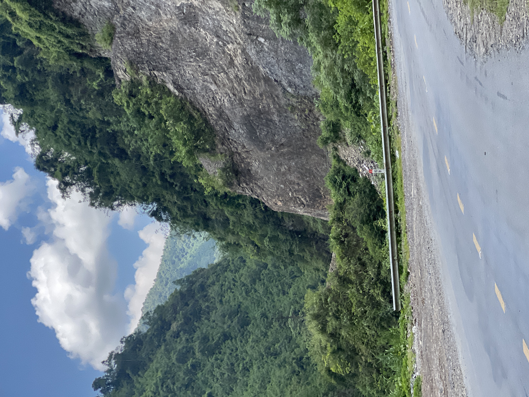 The image depicts a winding road cutting through a lush, mountainous landscape in Minh Tân Commune, Vietnam. On the right side of the road, a sheer, rocky cliff face rises dramatically, covered in patches of green vegetation. The left side of the road reveals a steep, densely forested mountainside, with layers of green foliage receding into the distance. The road itself is paved and appears to be a two-lane highway, marked with yellow lines. A metal guardrail runs along the edge of the road, separating it from the precipitous drop. The sky above is a bright blue, dotted with fluffy white clouds. The overall impression is one of natural beauty and rugged terrain.