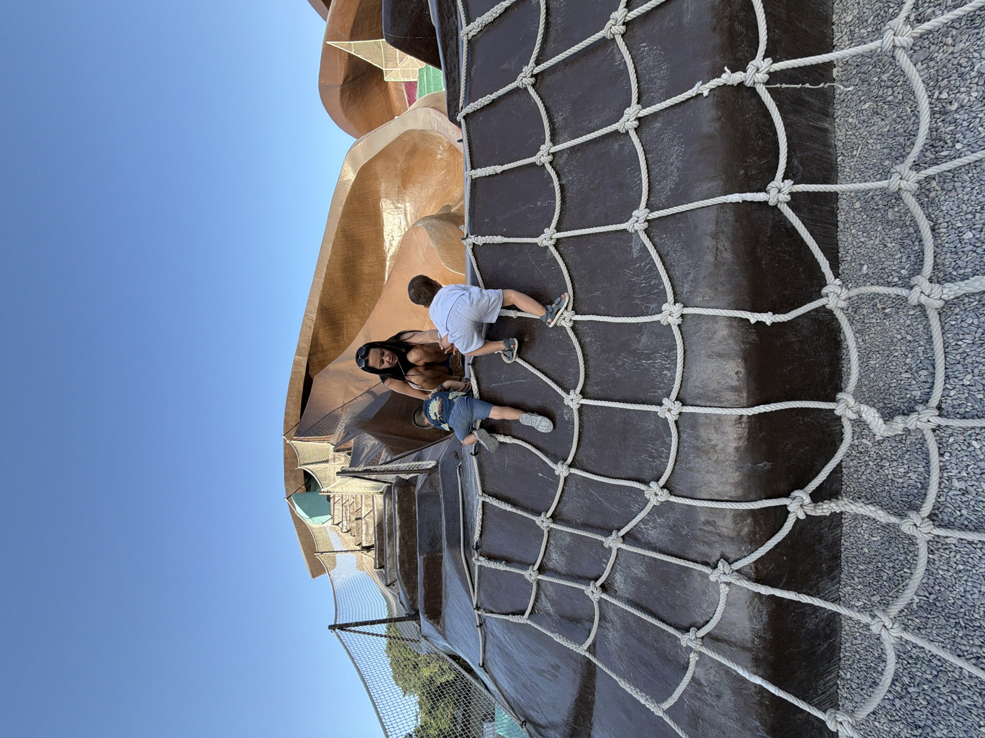 In this outdoor photograph taken in Valencia, Spain, two children are shown climbing on a playground structure. The image is oriented vertically. A young boy in a white t-shirt and grey shorts, wearing grey sandals, is actively climbing up a dark brown, curved surface with a net made of thick white rope. To his left and slightly below him, a woman with dark hair, wearing a bikini top and sunglasses, is also climbing. She is looking down towards the boy.
The playground structure appears to be made of a smooth, glossy material in shades of brown and copper, with some sections having a textured, pebble-like surface. The net is securely attached to the structure, providing a climbing aid. In the background, more of the elaborate playground can be seen, including curved slides and netting. The sky above is a clear, bright blue, indicating a sunny day, likely during daylight hours. The overall atmosphere seems to be one of active play and family enjoyment. No visible text is present in the image.