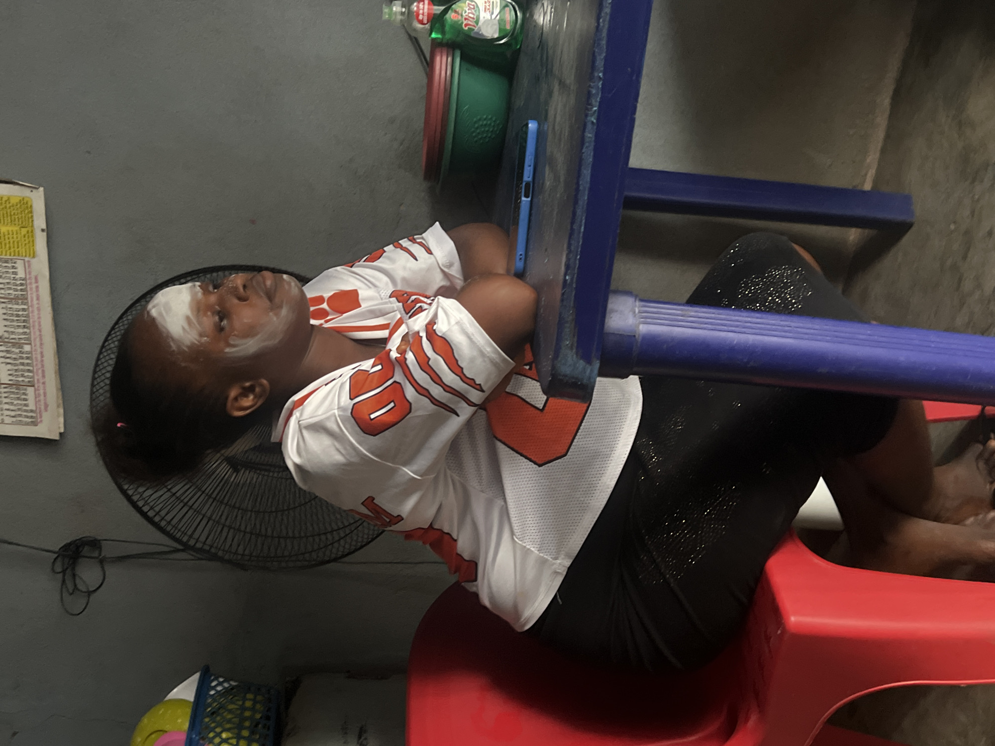 A young Black female with a white substance applied to her face, possibly a facial mask, sits on a red plastic chair. She is wearing a white jersey with orange accents and black pants. She is seated at a blue table, on which a blue mobile phone, two green plastic bowls, and a bottle of dish soap are present. In the background, a black standing fan is visible, along with a blue basket containing yellow and pink objects. The scene appears to be indoors, in a dimly lit room. The location is identified as Nkpor, Nigeria.