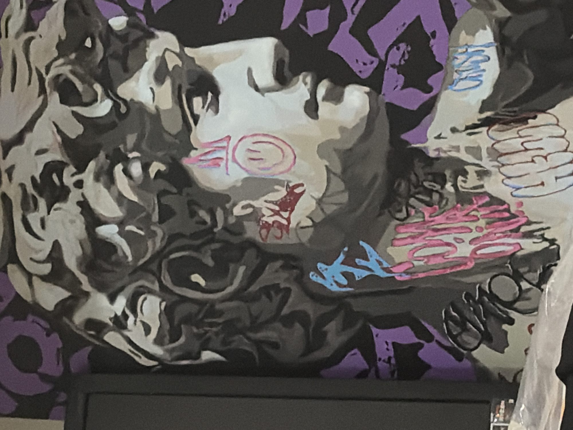 The image is a close-up of a mural depicting a classical bust, possibly Michelangelo's David, overlaid with graffiti. The bust is rendered in shades of grey and white, with stylized purple and black shapes in the background. Graffiti tags and symbols, including a smiley face and what appear to be various words in pink, blue, and white, are painted onto the face and chest of the bust. The overall style is a blend of classical art and contemporary street art. There is no indication of the location in the city of Chaikovsky, Russia, within the image itself.
