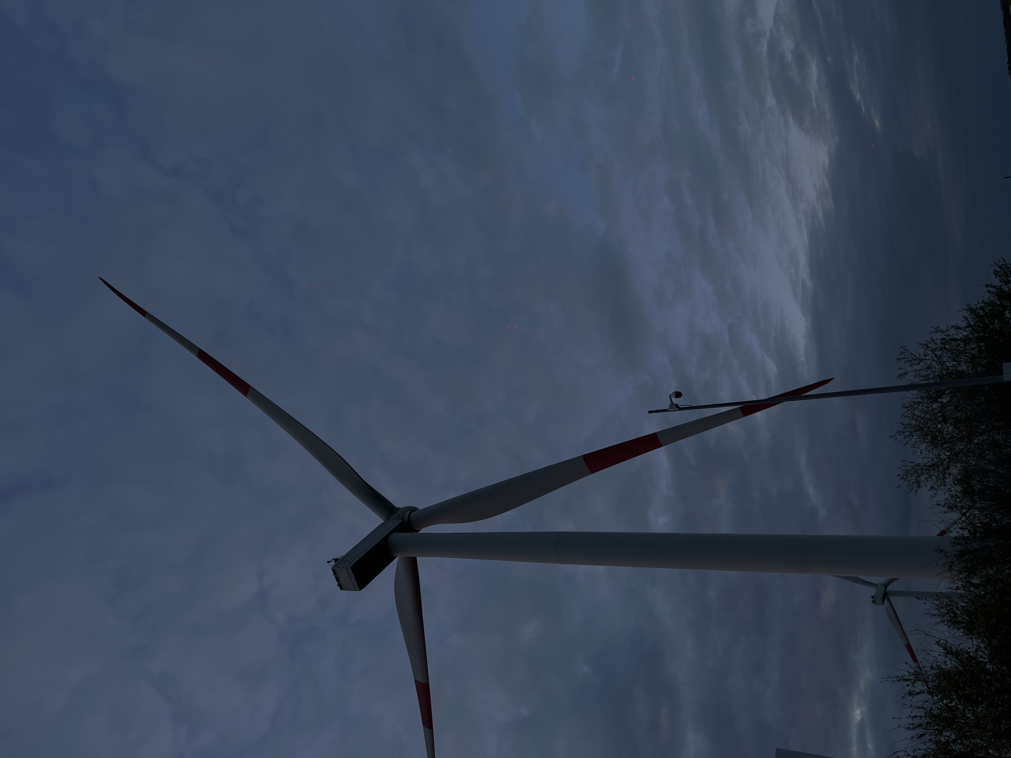 A low-angle shot depicts wind turbines against a cloudy, dark sky. The turbines are white with red tips on the blades. Some trees are visible to the right of the frame. The location, Port-Katon, Russia, is not visually verifiable.