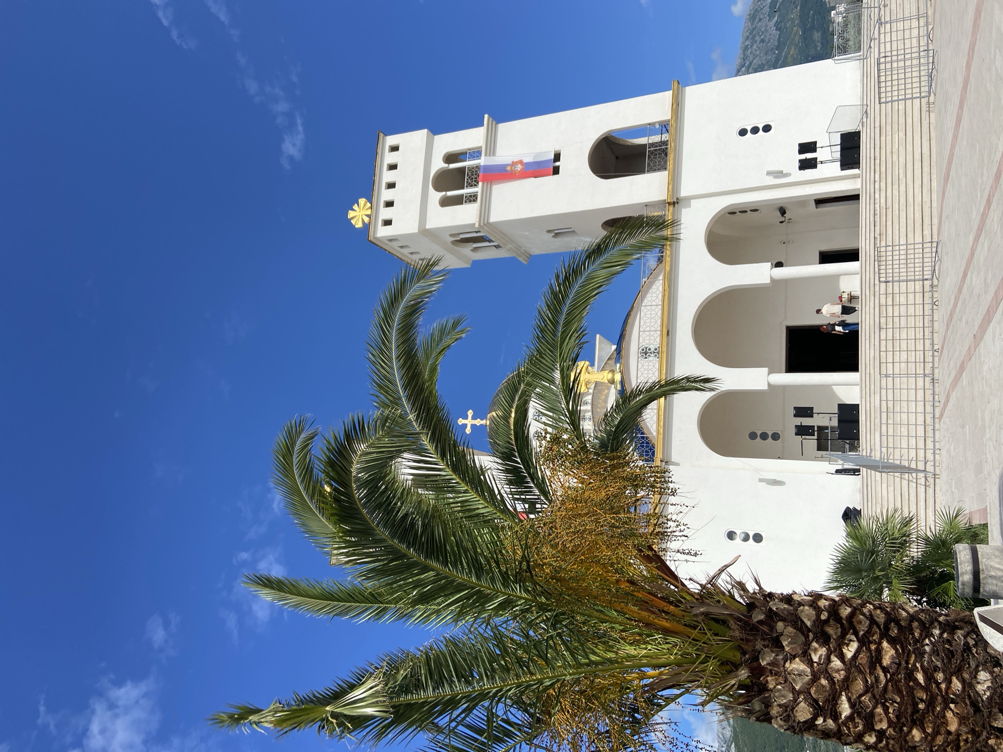 The image, when correctly oriented, depicts a modern white Orthodox church under a clear, vibrant blue sky. A large palm tree dominates the lower and central foreground, with its green fronds and yellow-brown seed clusters partially obscuring the view of the church.
The church features a prominent, rectangular bell tower adorned with a golden Orthodox cross at its apex. From an upper window of the bell tower, a flag of the Russian Federation (horizontal red, blue, and white stripes with a coat of arms in the center) is displayed. The building itself is characterized by clean white facades, multiple arched openings, and decorative grilles on some windows. Another golden Orthodox cross is visible on a lower dome or roof section.
Two individuals are standing near an arched entrance on the right side of the church. The ground in front of the building appears paved or tiled, with some metal barriers or railings. In the far background, on the left, a portion of a mountain range is visible beneath the clear sky. The scene is brightly lit by sunlight, suggesting a clear day. No discernible text is visible on the building or its surroundings, apart from the coat of arms on the flag.