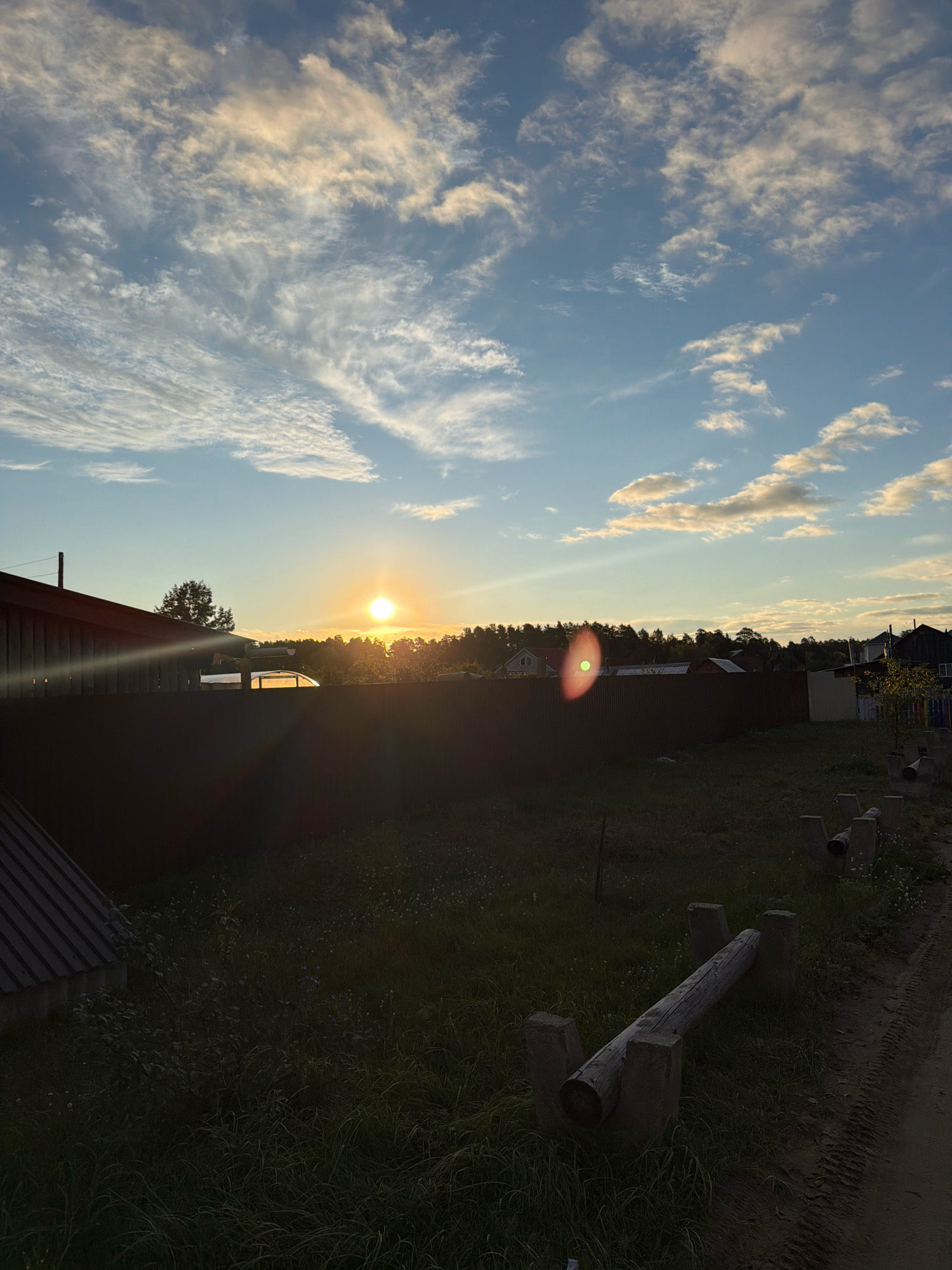 The image captures a sunset over a rural or suburban area, likely in Chaikovsky, Russia. The sun is low on the horizon, casting a warm glow and creating lens flare effects. The sky is partly cloudy with wispy cirrus clouds and larger, more defined cumulus clouds, illuminated by the setting sun.
In the foreground, a dark, corrugated metal fence runs horizontally across the frame. To the left, the corner of a wooden building with a sloping roof is visible. The ground in front of the fence is covered in green grass with scattered wildflowers. Further into the scene, beyond the fence, a line of trees forms a dark silhouette against the bright sky. Several structures, possibly houses or sheds, are visible nestled among the trees.
On the right side of the image, a dirt path runs alongside the grassy area. Several wooden logs supported by concrete blocks are positioned on the grass, suggesting an outdoor obstacle course or exercise equipment. The overall impression is a peaceful, late-evening rural landscape.