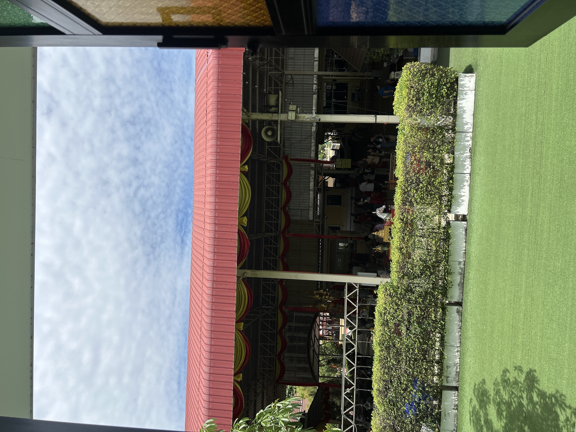 This image appears to be taken from within a building, looking out through an open window or doorway towards an outdoor covered area and a grassy lawn. The covered area has a red roof with yellow and red fabric draped along the edge. Underneath the roof, there are people gathered, some standing and others seated. There are also statues and decorative elements visible, suggesting a temple or a place of worship. A white metal fence separates the covered area from a manicured green hedge. Beyond the hedge, a bright green artificial lawn is visible, with shadows of trees cast on it. The sky above is partly cloudy with scattered white clouds against a blue sky. The foreground is framed by what appears to be the edge of a window or door with a stained-glass element on the right side. The presence of a statue of a horse-like figure in front of the hedge might indicate a festival or a special event. Based on the context of a temple-like structure and the surrounding environment, this scene is likely located in a tropical or sub-tropical region, possibly in Thai Ko Subdistrict, Thailand, given the information provided.