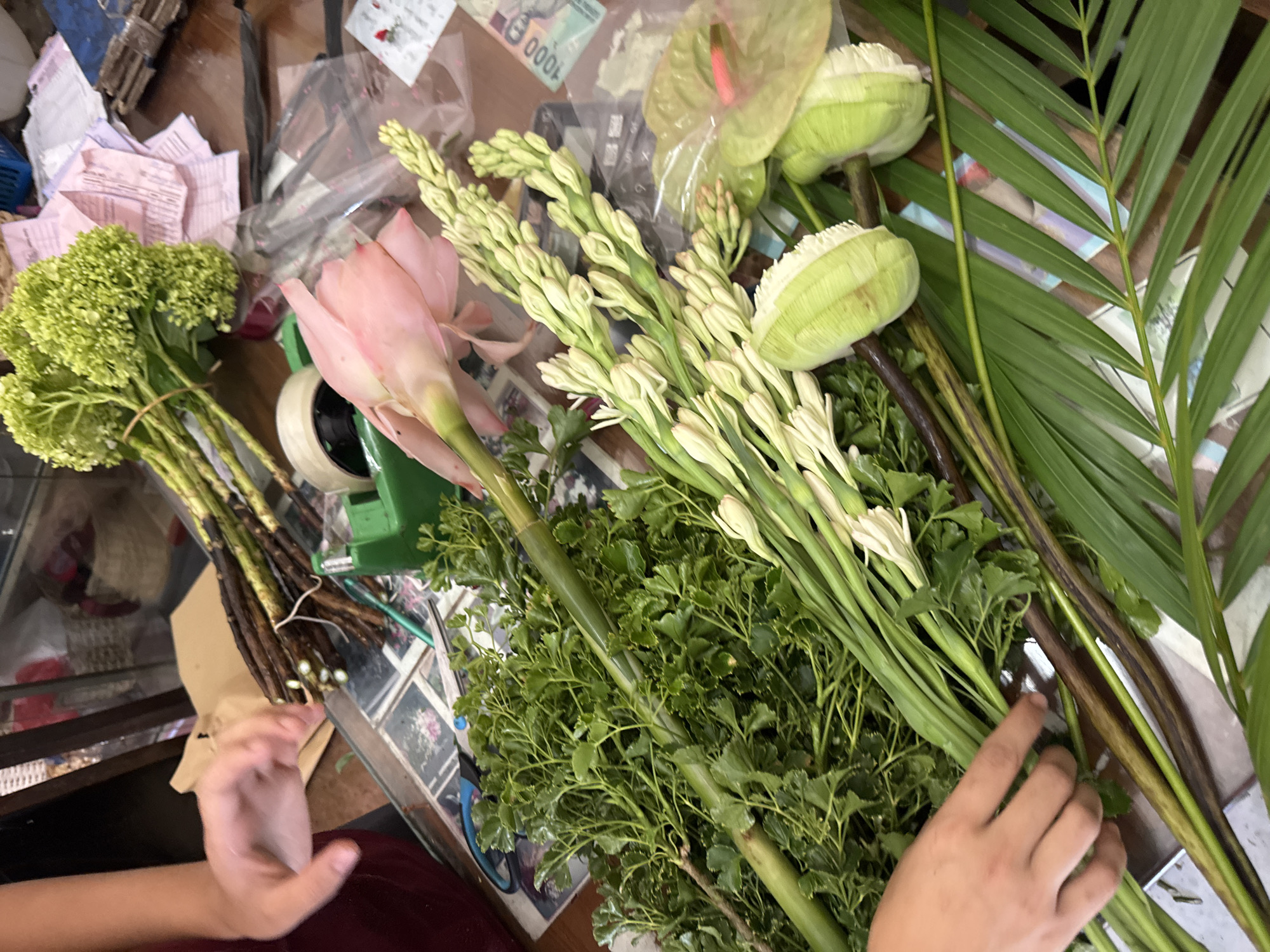 The image shows a collection of flowers and greenery on a table. Visible are green hydrangeas, pink ginger lily, white tuberoses, light green anthurium, and palm fronds. A roll of tape, scissors, and other tools are also present on the table. A person's hands are visible holding the flowers. In the background, Indonesian currency can be seen. The image was likely taken in North Kuta, Indonesia.