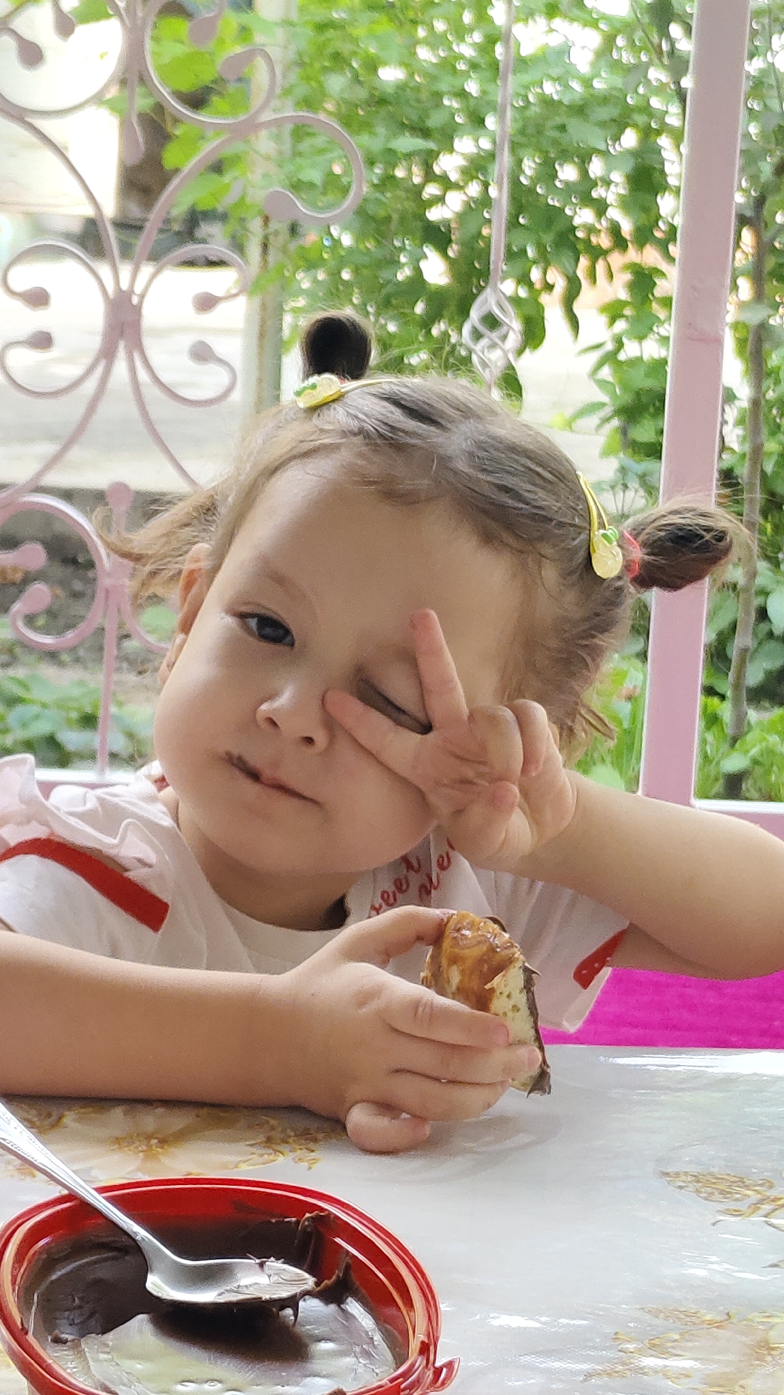 A young girl with chocolate smudged on her lips sits at a table outdoors, holding a piece of pastry and making a peace sign with her fingers. She has her hair pulled up in two small buns, adorned with yellow clips. A red bowl of chocolate spread and a spoon are visible on the table in front of her. The background consists of greenery and a pink decorative trellis, suggesting a garden setting, possibly in the city of Akkurgan, Uzbekistan.