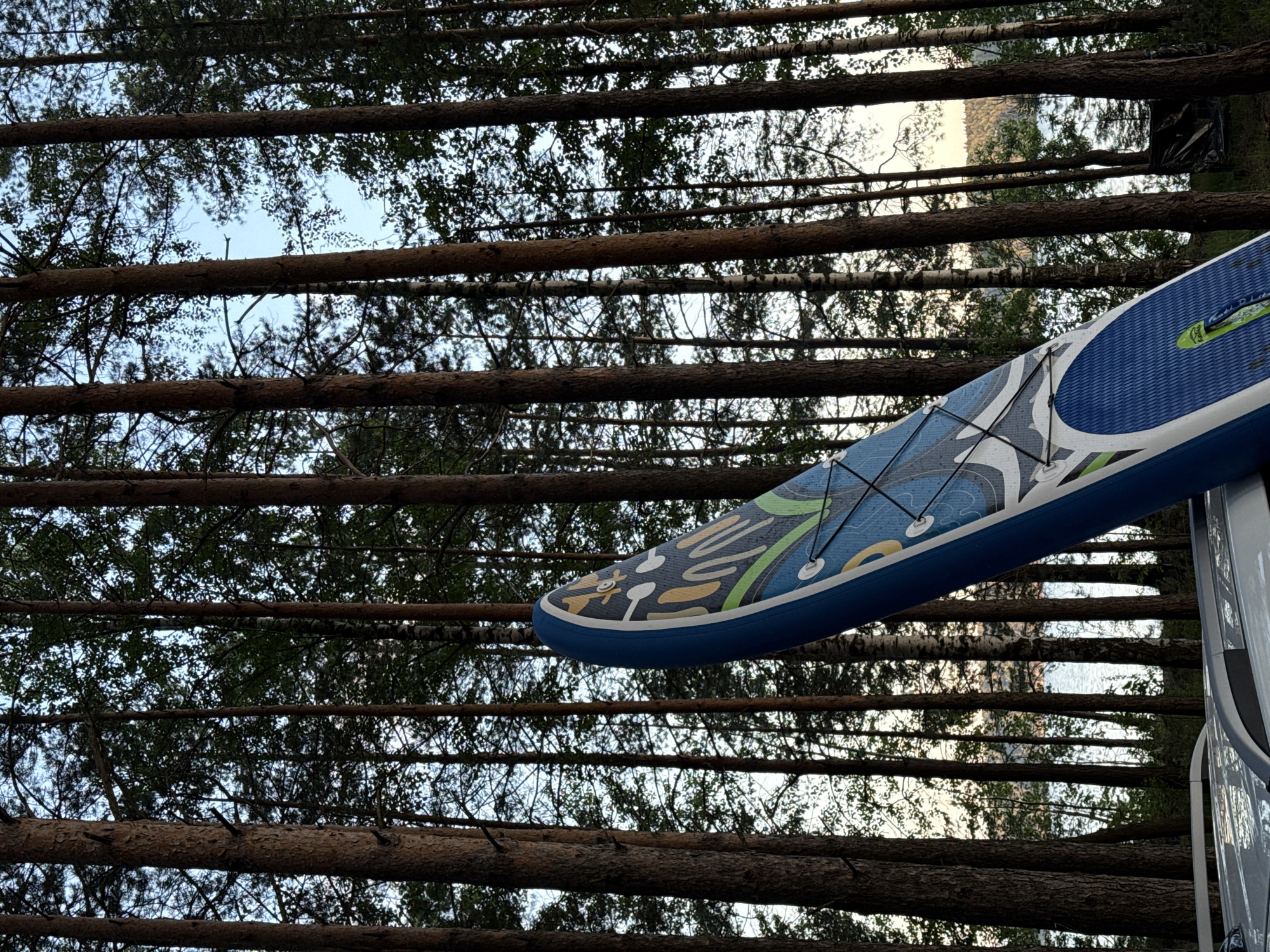 An angled shot features the front of a blue and white paddleboard leaning on the roof of a car, set against a backdrop of tall pine trees. The paddleboard displays a colorful abstract design. Beyond the trees, a body of water, possibly a lake, is visible with a distant tree-lined shore. The scene is set in a forested area near a body of water, with the location identified as Gora (Khokhlovskoe S/P), Russia. No people are present in the frame. The lighting suggests it is either early morning or late afternoon.
