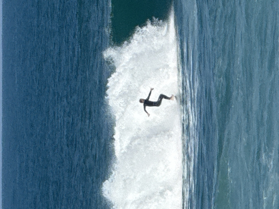 A lone individual, clad in a dark full-body wetsuit, is depicted actively surfing on a white surfboard. The person is positioned mid-action on the face of a breaking white-water wave, with both arms extended horizontally for balance. Bare feet are visible on the surfboard. The immediate water around the surfer is a lighter blue, transitioning to a darker, deep blue ocean extending towards the horizon in the background. This scene is situated in the Mediterranean Sea, off the coast of Tel-Aviv, Israel.