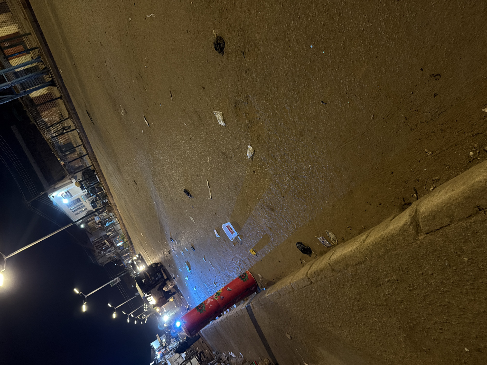 The image depicts a street scene at night in Bukuru, Nigeria. The road surface is wet and appears to be littered with debris, including plastic bags and other trash. A red, cylindrical object with a floral pattern stands near the curb on the left side of the frame, illuminated by a blue light source from the left. In the background, several vehicles are visible, including what appears to be a three-wheeled auto-rickshaw. Buildings line the street, some of which have lights on. Streetlights illuminate the scene from above. The overall impression is of a somewhat unkempt urban environment at night.
