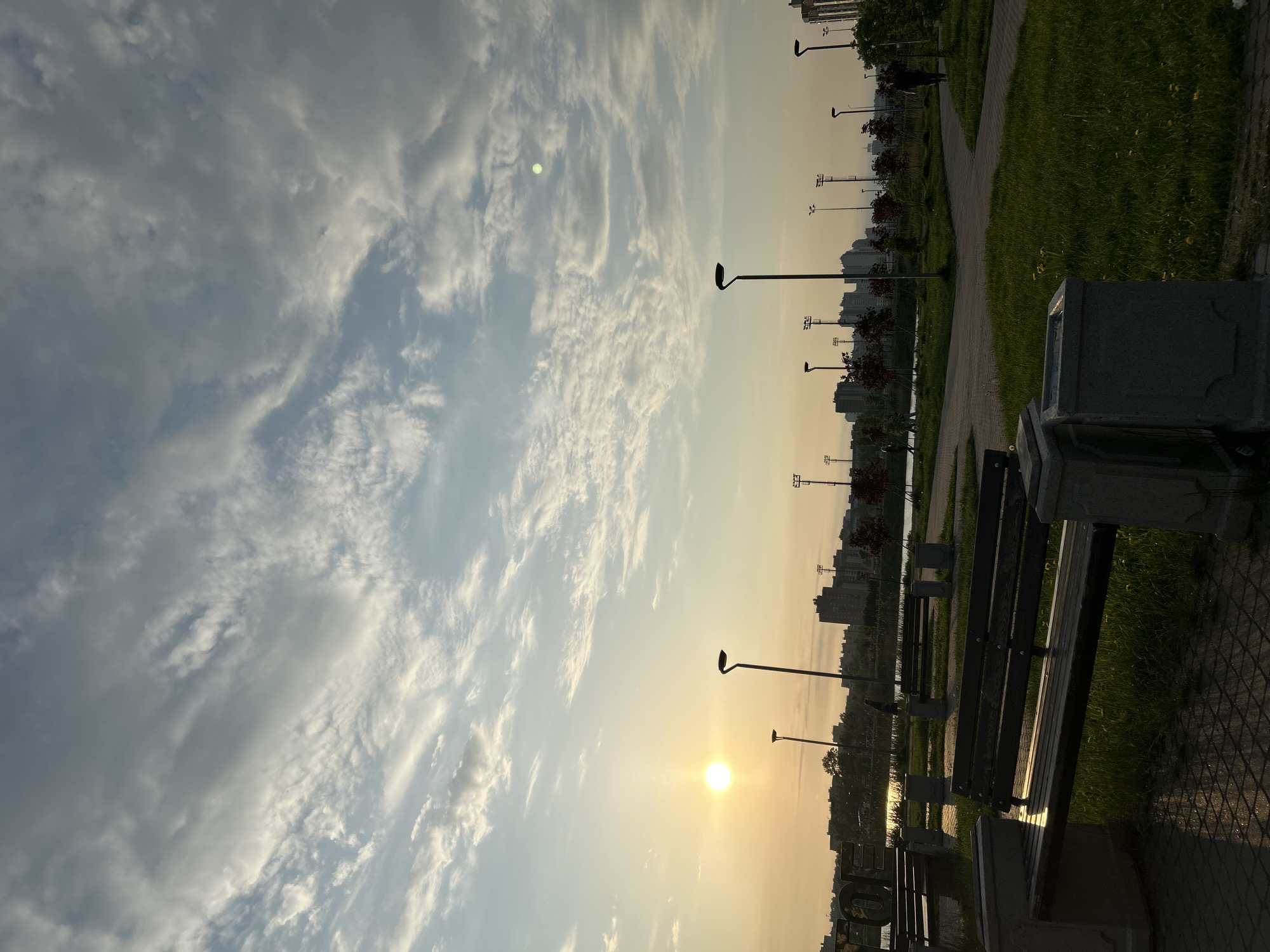 The image shows a park scene at sunset in Saint Petersburg, Russia. A row of benches and trash receptacles lines a paved walkway that runs along a water feature, possibly a canal or river. Lampposts are spaced along the walkway. Buildings are visible in the background, indicating an urban setting. The sky is filled with clouds. The sun is low on the horizon.