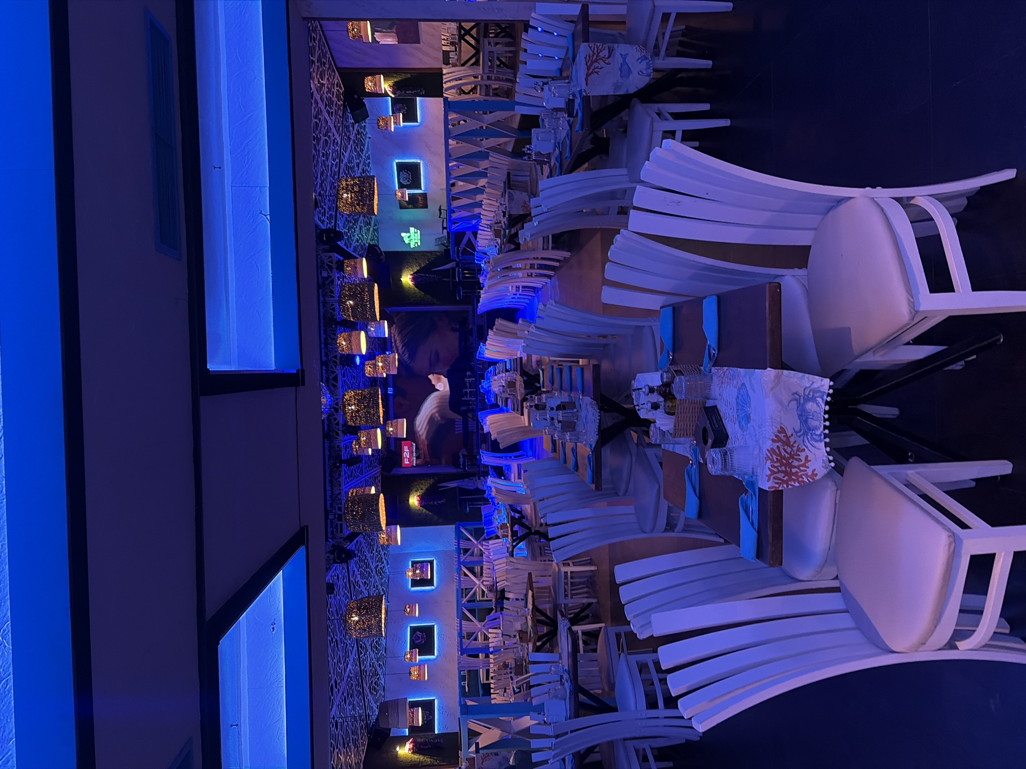 The image depicts the interior of a restaurant. The dominant color is blue, likely from artificial lighting. White chairs with curved backs are arranged around wooden tables. Table settings with glasses and decorative cloths are visible. Ornate, hanging lamps adorn the ceiling, and patterned walls add to the decorative scheme. A large photograph is on the back wall. The location is Plovdiv, Bulgaria.