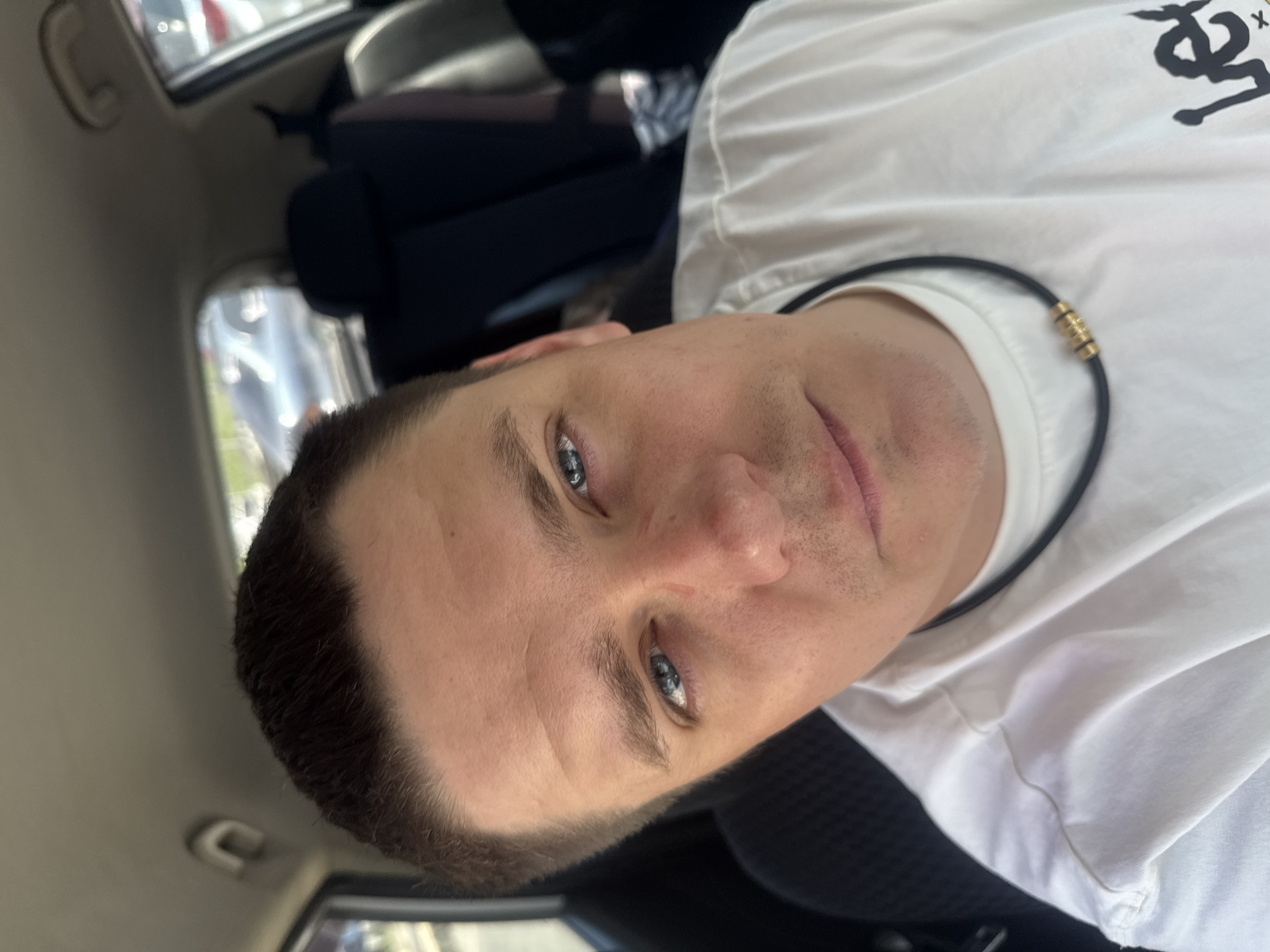 A man with short brown hair and blue eyes is in the driver's seat of a car. He is wearing a white t-shirt with a black necklace that has gold beads. The car appears to be parked in an outdoor location, possibly a parking lot, with other vehicles visible in the background through the car windows. The lighting suggests it is daytime.