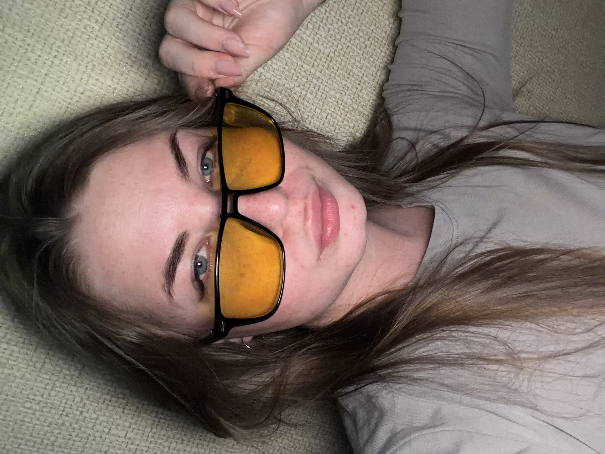 A fair-skinned young woman with light brown hair is lying down, possibly on a sofa, with her head tilted slightly to the right. She is wearing a light grey t-shirt and black-framed glasses with yellow lenses. Her left hand is near the glasses, and her gaze is directed toward the camera. The background shows a beige textured fabric, likely the sofa or bedding. The location is not evident in the image, and there is no evidence that it is in Chaikovsky, Russia.