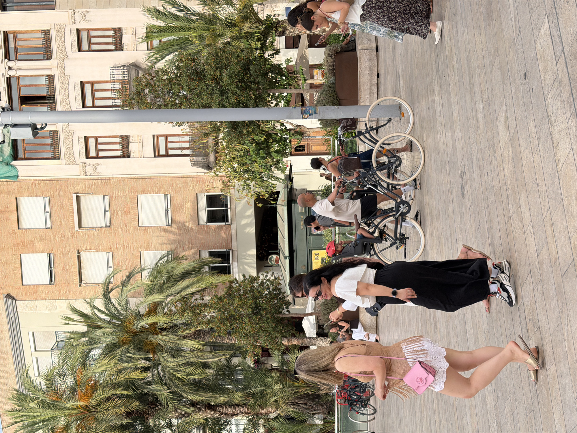 A street scene in an urban environment features a group of people in the foreground walking and standing amidst palm trees and buildings. In the center, a woman in a white off-the-shoulder top and black pants is smiling and looking to her right, holding a mobile phone. To her left, another woman in a crochet-like brown top and white fringed skirt is walking away from the camera, carrying a pink purse. Several bicycles are present, with two parked in the middle of the frame and another visible on the left. In the background, people are seen walking and interacting. The buildings are multi-story structures with brick and cream-colored facades, featuring balconies and windows. Palm trees and other greenery are interspersed throughout the scene. The ground is paved with light-colored rectangular tiles. The lighting suggests it is daytime, likely late afternoon given the shadows.