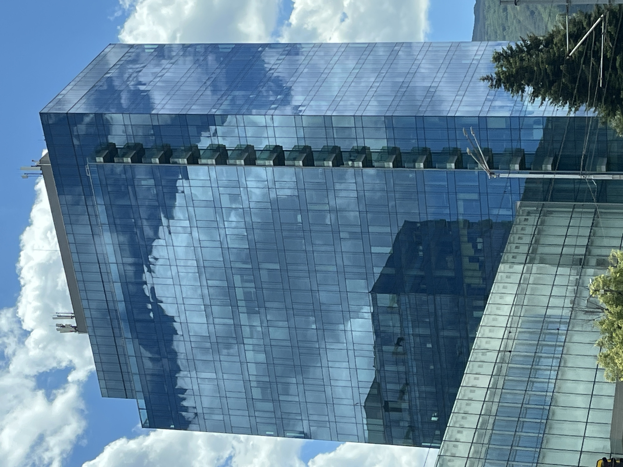 A tall, modern glass skyscraper dominates the center of the frame, its blue-tinted facade reflecting the sky and clouds. The building has a rectangular footprint and a series of balconies that project from one side. The top of the building features communication antennas. In the foreground, a lower, horizontally oriented glass building is partially visible, with rows of windows. To the right of the skyscraper, a mature evergreen tree stands tall, with a mountain range visible in the background. A street light and utility wires are also present in the scene. The sky is a clear blue, dotted with fluffy white clouds.