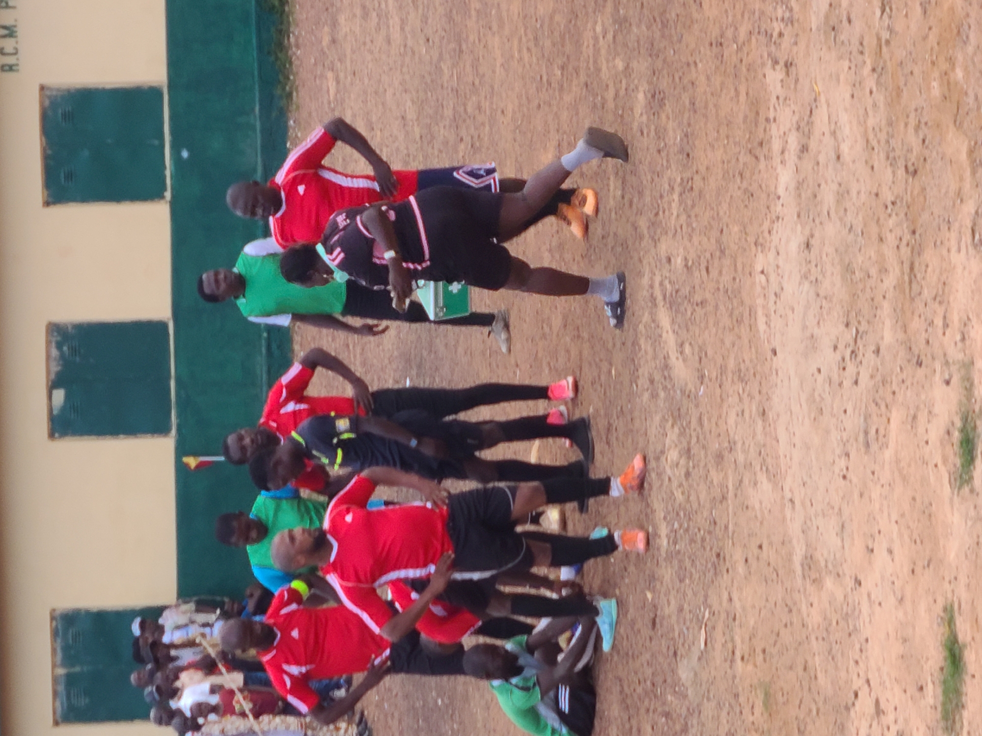 A group of men in soccer uniforms and casual attire are gathered on a dirt field. The men in uniform appear to be players and possibly a referee. One man in a red jersey with white trim is in the foreground, facing away from the camera. Another man in a similar red jersey is to his left. A man in a green and white jersey stands behind them. To the right, a woman in a black and pink uniform is holding a green box. In the background, a building with green window frames and the partially visible text "R.C.M. P" is seen. The ground is uneven dirt with some patches of dry grass. The scene suggests a casual soccer match or practice is taking place in an outdoor urban setting. The presence of the building with "R.C.M. P" might indicate a school or community center in Jos, Nigeria, given the visual context.