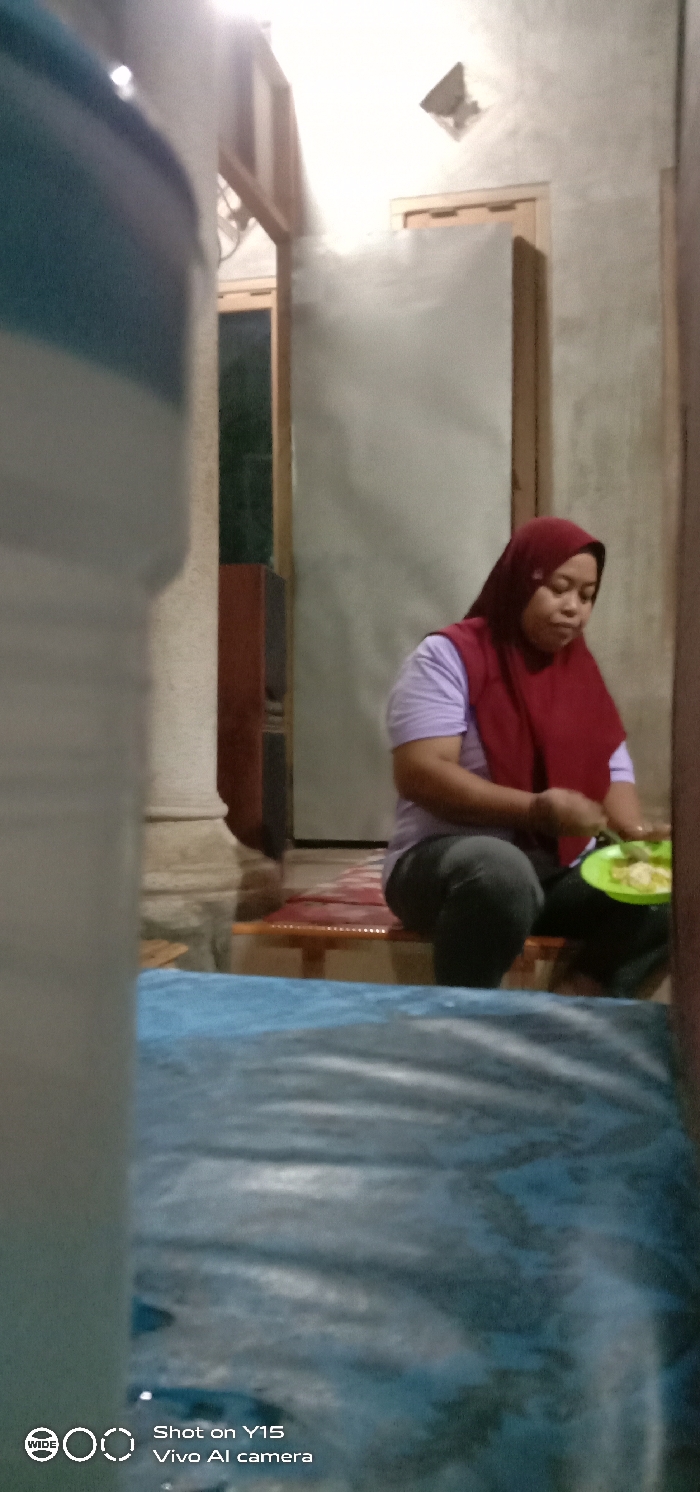 The image depicts an indoor or semi-enclosed residential area in Jabungwetan, Indonesia, illuminated by artificial light. An adult female, of apparent Southeast Asian descent, is seated on a low wooden bench covered with a reddish-brown mat. She is wearing a light purple short-sleeved top, dark trousers, and a maroon hijab that covers her head and chest. She holds a bright green plate containing light-colored food, which she appears to be eating or preparing to eat, her gaze directed downwards.
To the left in the foreground, a large, blurry, white and blue-striped cylindrical object partially obstructs the view. The floor in the immediate foreground is covered by a patterned blue-grey fabric or mat. The background features light-colored walls, possibly stucco or painted concrete, with various wooden architectural elements, including door frames and structural beams. A dark brown cabinet or shelving unit is partially visible on the left side of the background. A small, square wall vent is visible high on the right background wall. The scene's lighting is even, suggesting an overhead light source. A "Shot on Y15 Vivo AI camera" watermark is present at the bottom left.