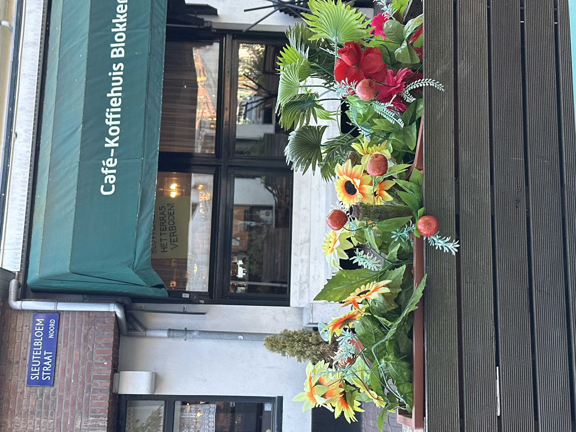 The image shows a street view in Amsterdam, Netherlands, featuring a cafe named "Café-Koffiehuis Blokker". A green awning covers the cafe's entrance. A street sign is visible, reading "Sleutelbloem Straat, Noord." Flower arrangements decorate the exterior of a building. The weather appears fair.