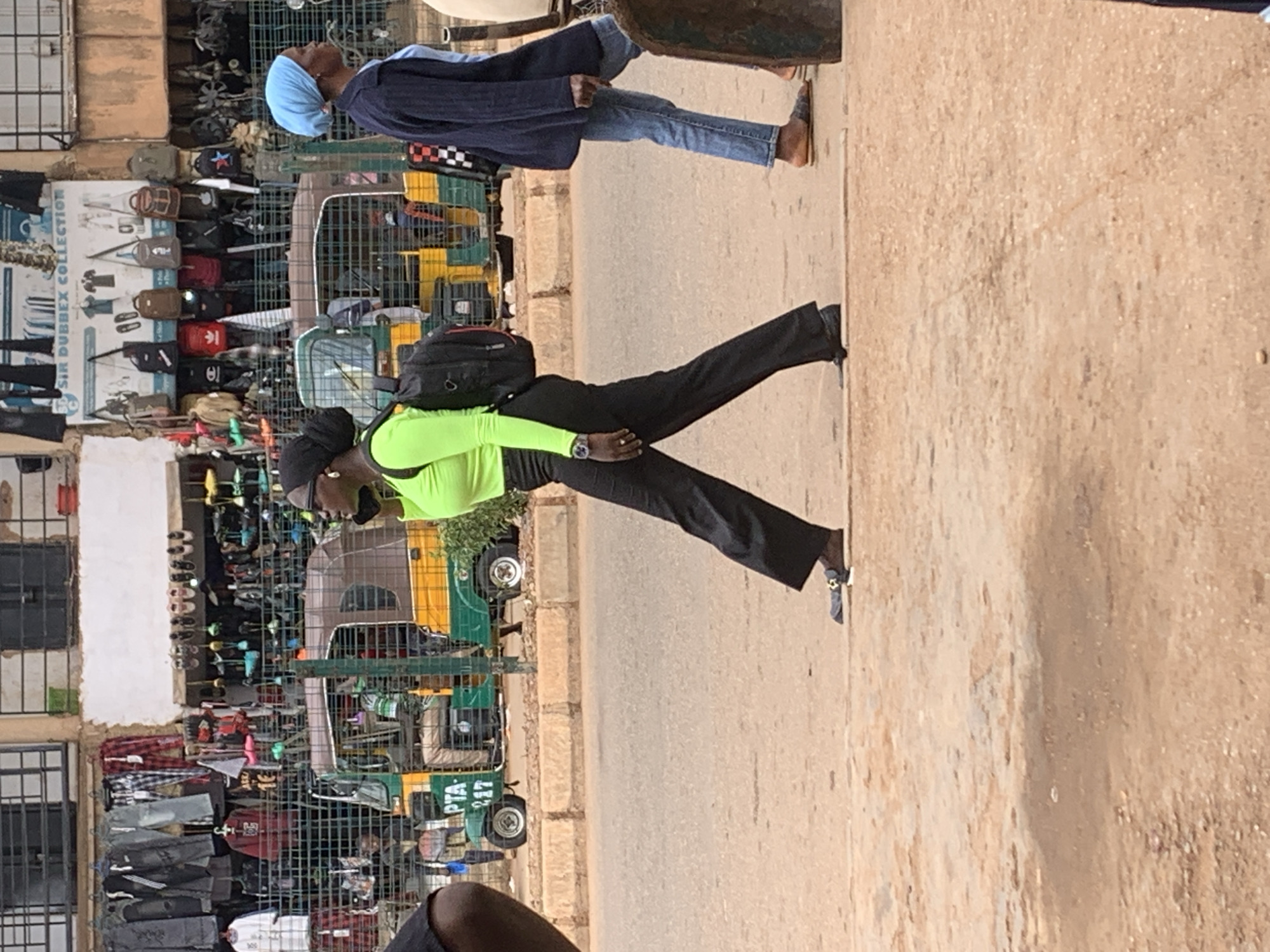 The image shows two people in Bukuru, Nigeria. The subject in the foreground is wearing a bright green long-sleeved shirt and black pants, with a black backpack. They appear to be standing or posing, with one leg extended. A watch is visible on their left wrist. Behind them, another person is seen wearing a blue head covering and a dark blue jacket, with blue jeans. They are walking or standing next to a cart.
The scene appears to be a market or roadside stall area. Racks of clothing and bags are visible behind the foreground subject, with a variety of items displayed. A green and yellow tricycle-like vehicle is also present, along with other carts or stalls. The ground is a dirt or asphalt road. The lighting suggests it is daytime, likely sunny.
Visible text on the tricycle reads "PTA 247". There is also a poster on the left side of the image with text that includes "SIR DUBBEX COLLECTION". The overall atmosphere appears to be a busy outdoor market setting.