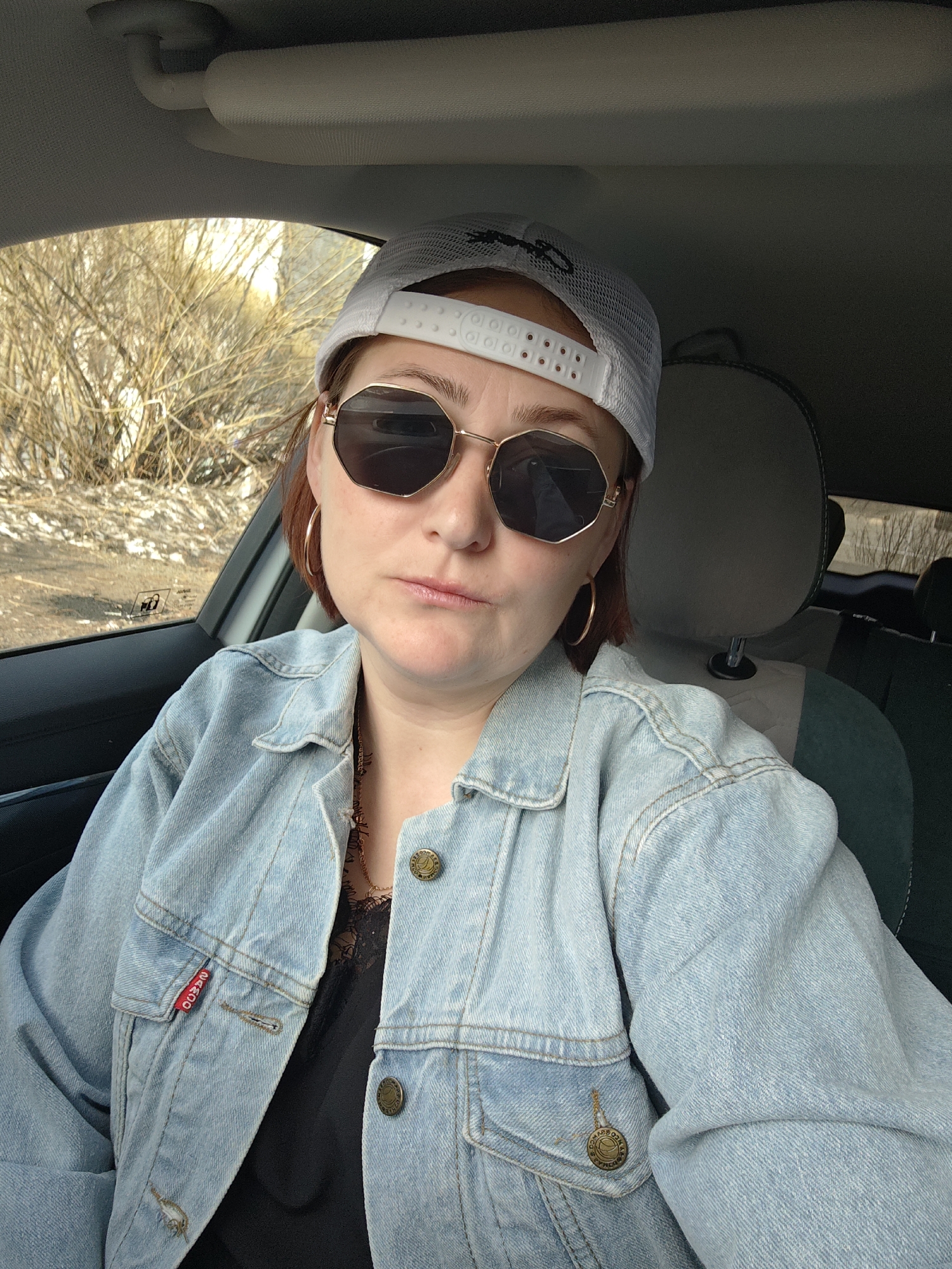 A woman is sitting in a car in Markovskii, Russia. She is wearing a light blue denim jacket, a black top, a white baseball cap, and gold sunglasses. She has gold hoop earrings and a necklace. The car is parked near a tree.
