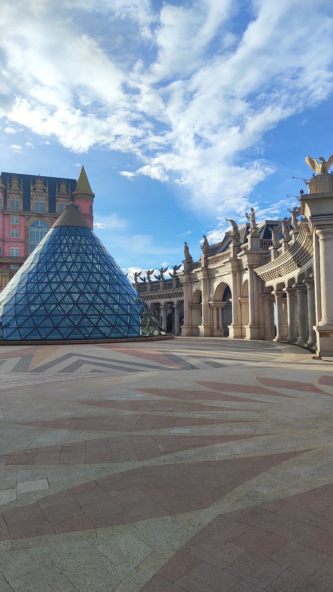 This image captures an outdoor plaza in front of a building complex in Da Nang, Vietnam. Dominating the left side of the frame is a large, translucent glass pyramid with a metallic frame, its surface reflecting the blue sky and clouds. Behind the pyramid, a pink building with dark blue roof gables and yellow spires is partially visible. To the right, a grand, arched colonnade stretches across the scene, adorned with statues and intricate architectural details. The ground is paved with large square tiles in patterns of pinkish-red and beige. The sky above is a bright blue, populated by scattered white clouds. There are no people visible in the image. The architectural style suggests a fantasy or theme park setting, consistent with attractions like Sun World Ba Na Hills in Da Nang.