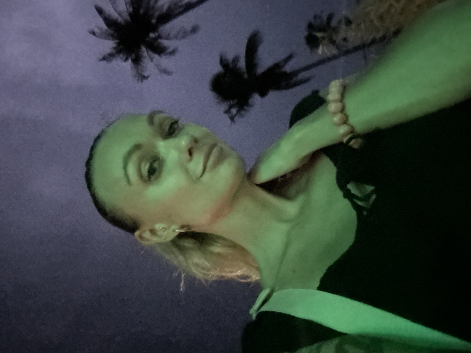 A low-angle selfie photograph taken at night in Mũi Né, Vietnam, features a light-skinned female posing against a backdrop of palm trees and a dark purple sky. The woman is fair-skinned with blonde hair pulled back. She is wearing a black top with a white collar and a pink beaded bracelet on her right wrist. Her right hand is raised and touching her neck. The overall scene has a green cast. Palm trees are visible silhouetted against the night sky.