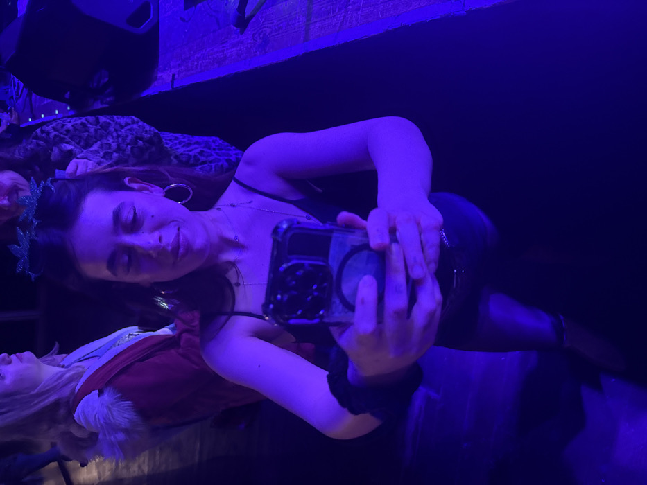 A woman in a black tank top with a green leaf crown and hoop earrings holds a phone up to take a selfie. She is smiling slightly and looking at the screen. She wears multiple necklaces and a black scrunchie around her wrist. In the background, another person is laying in a Santa outfit next to a person in leopard print. The scene is lit with blue lighting.