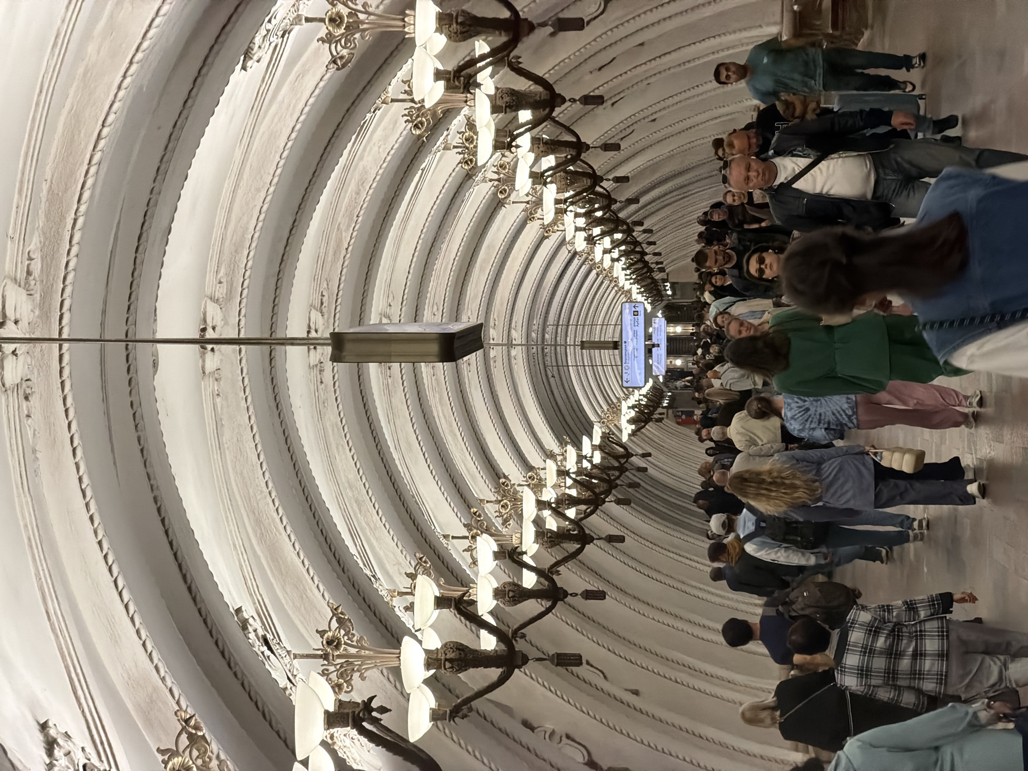 The image shows a subway station in Moscow, Russia, oriented with the ceiling at the top and the floor to the right. The ceiling is curved with decorative ribs and ornate lighting fixtures. A crowd of people are walking along the station platform. Signs are visible down the length of the station.