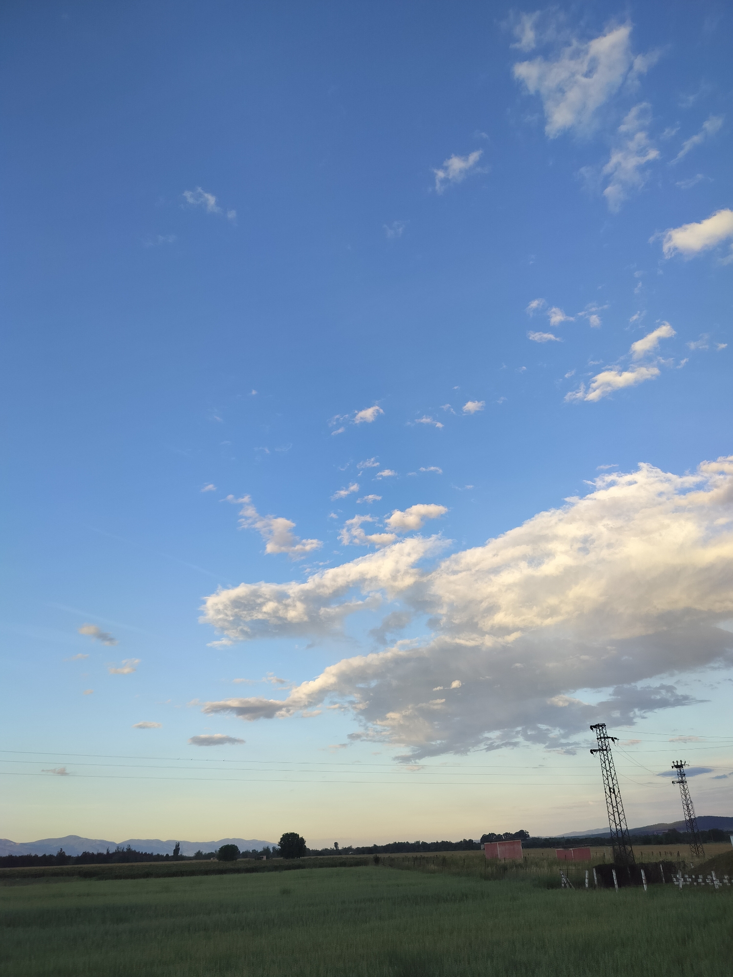 The image shows a wide, rural landscape under a bright blue sky with scattered white clouds. In the foreground, a green field extends across the bottom half of the frame. In the mid-ground, a line of trees and some low buildings are visible, along with two tall electricity pylons supporting power lines that stretch horizontally across the scene. In the distance, a range of mountains forms the horizon line. The overall impression is one of a peaceful, open, and natural environment.