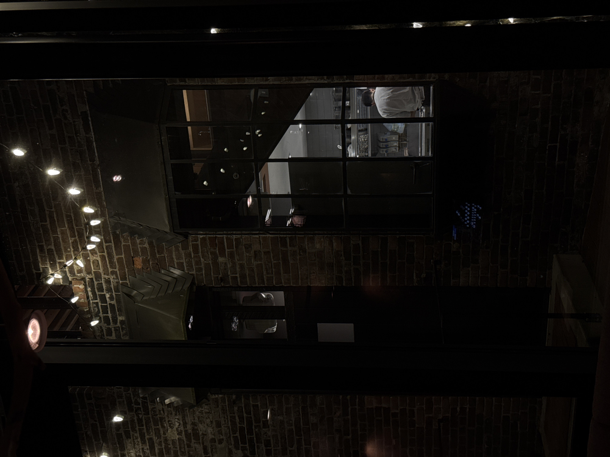 The image depicts an exterior view at night, potentially in Vancouver, Canada, given the prevalence of brick architecture in older parts of the city. Focus is given to a brick wall structure with two visible windows. The upper window provides a partial view into what appears to be a commercial kitchen, showing stainless steel appliances and a person wearing a white chef's coat. The lower window shows blurry figures in a room. String lights hang across the top left of the frame, partially obscured by the architectural structure, adding ambient light to the scene. The scene is generally dark. The foreground is composed of black metal framing. The architectural style and lighting suggest a restaurant or bar setting.