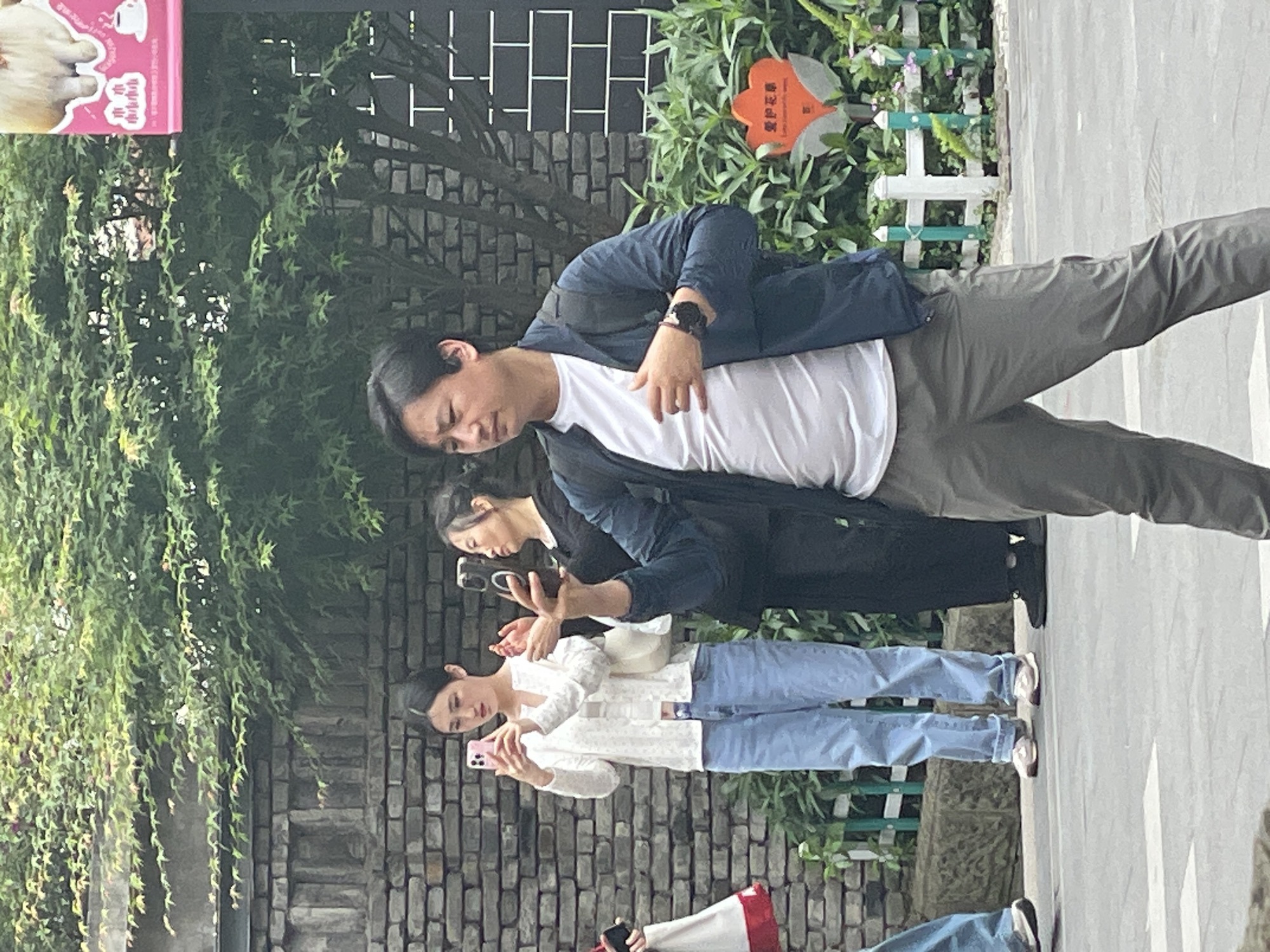 A group of at least four people are on a city street with stone or brick walls and green plants in Yuzhong District, China. The first person in the line, likely a man, wears grey pants, a white shirt, and a dark jacket. The other three people following are also looking at their phones. The first person holds their hand on their stomach and looks downwards. The people seem to be tourists.